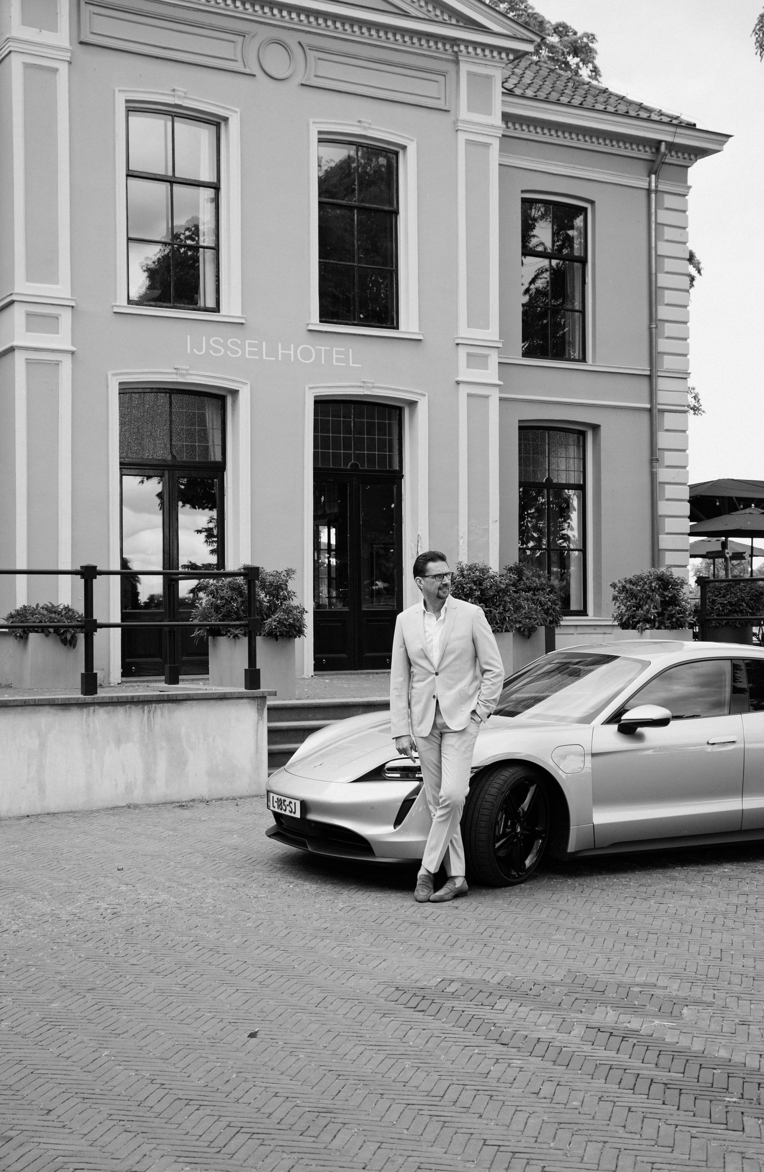 Groom in a light suit standing beside a Porsche in front of the IJsselhotel building.