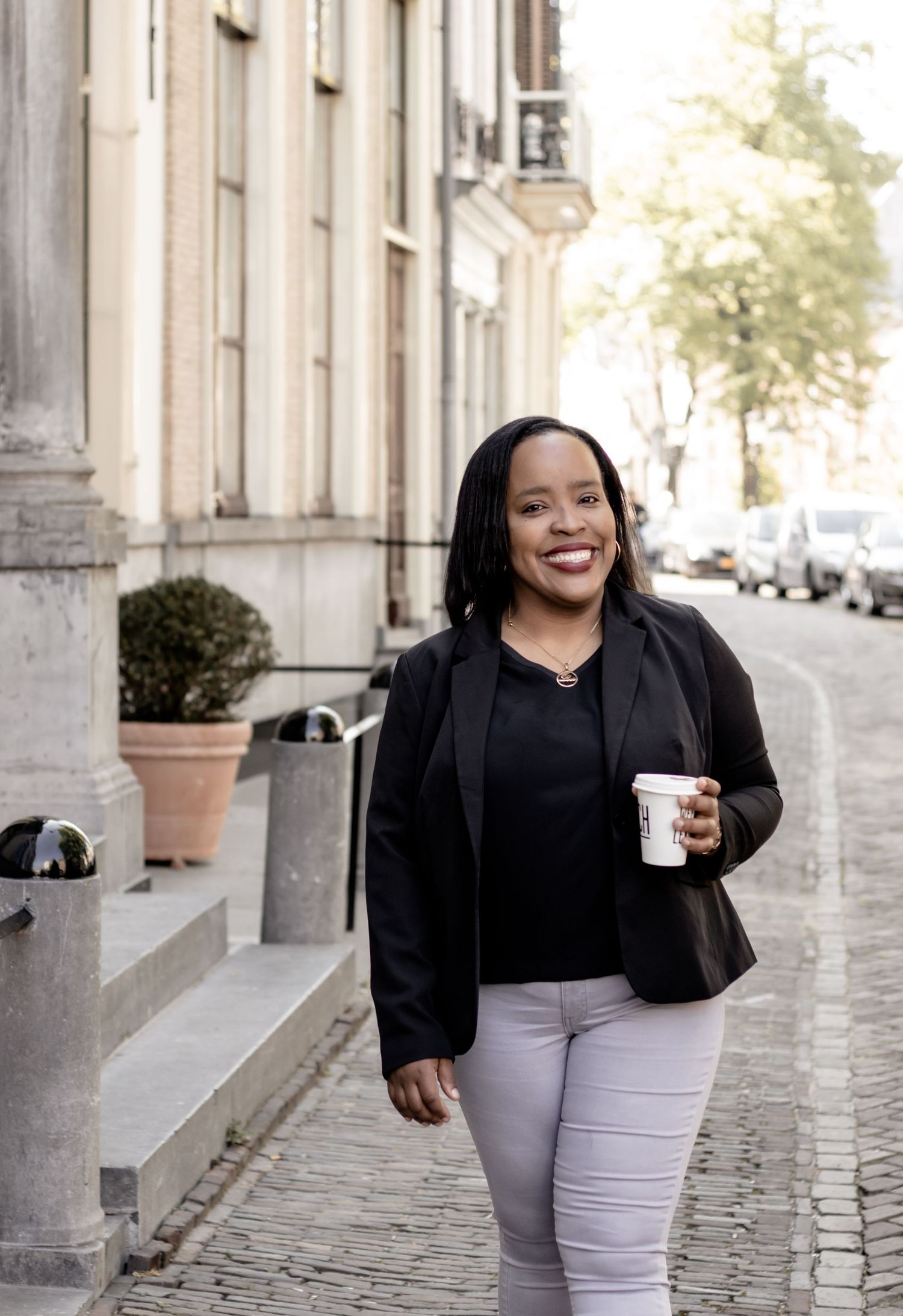 Smiling Black woman in black blazer walking on a cobblestone street holding a coffee cup.