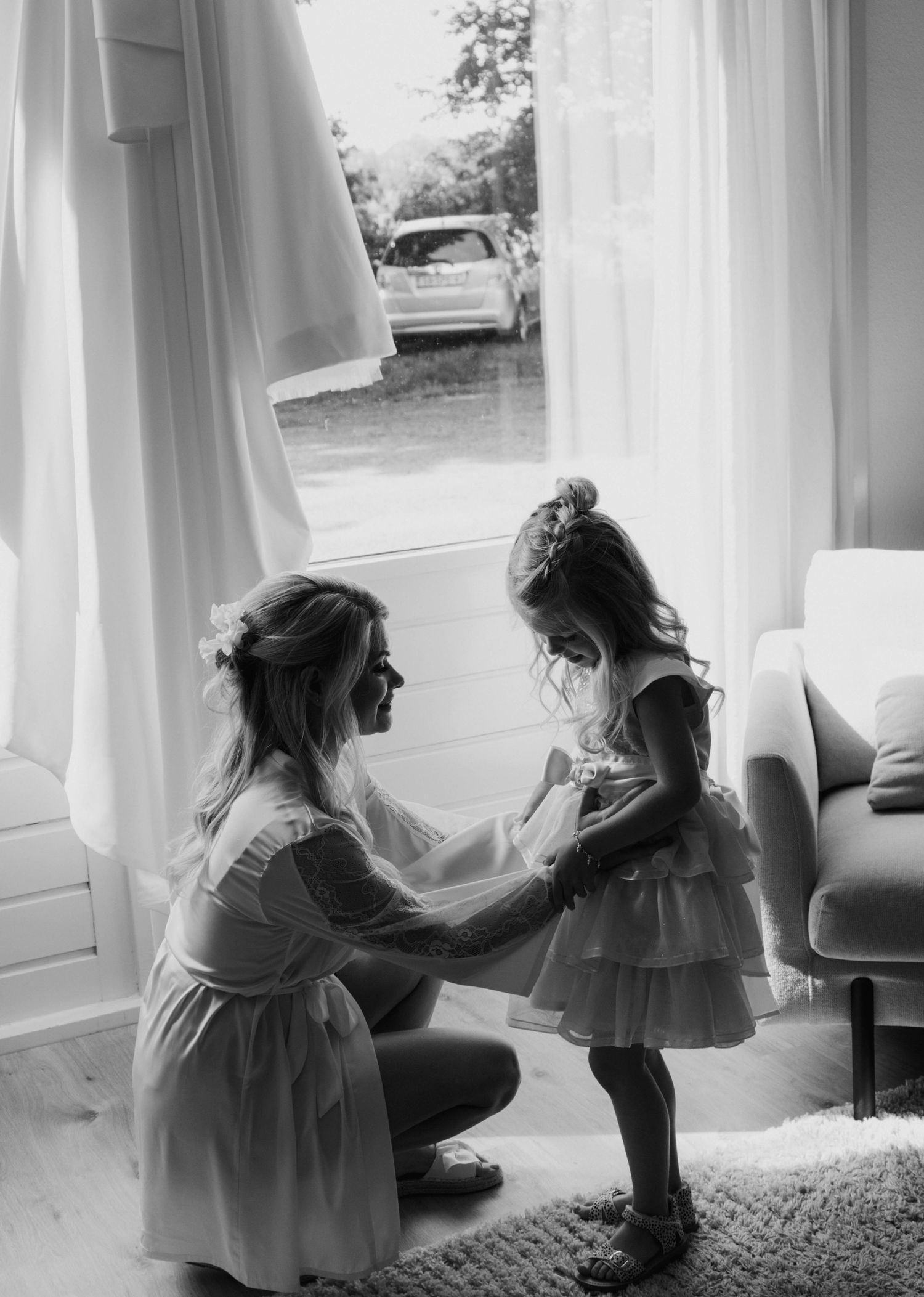 Black and white photo of a woman kneeling to adjust a young girls dress near a bright window.