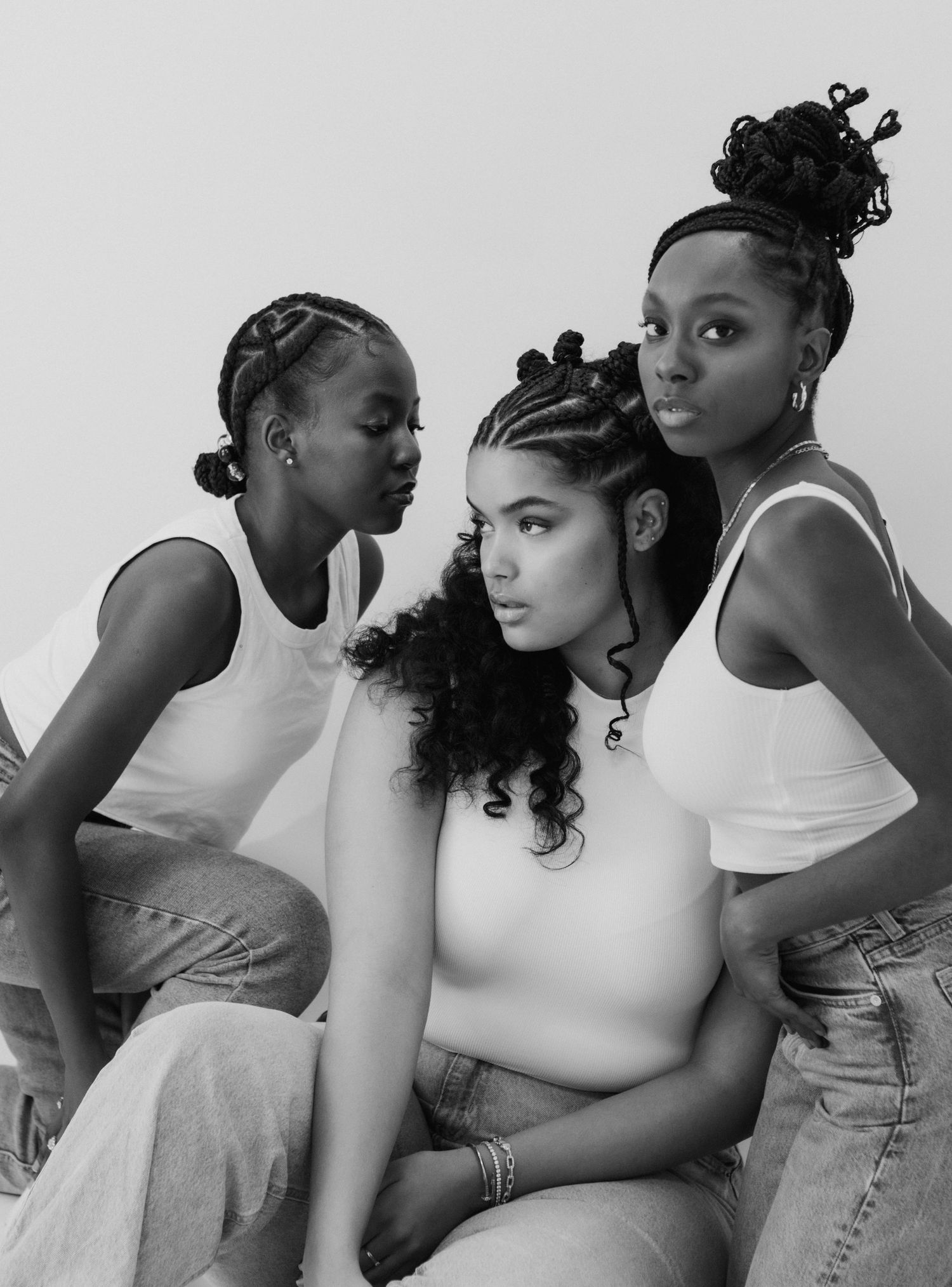 Three young Black women posing together in white tops and jeans in a black and white photo.