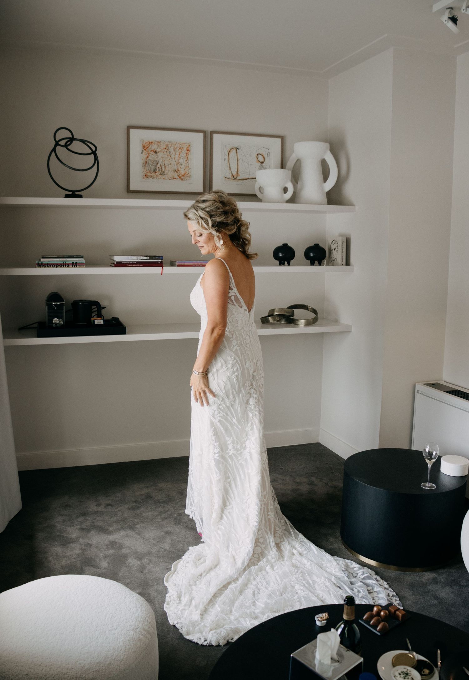 Bride in elegant lace wedding gown with train standing in a stylish hotel room before her wedding.