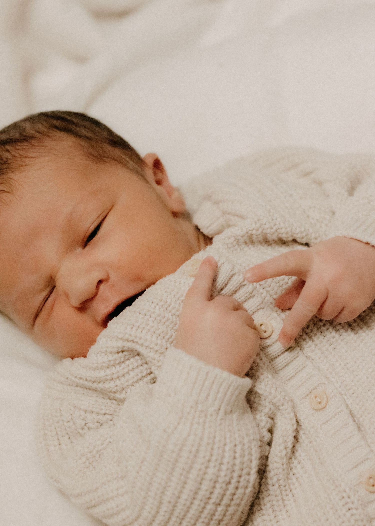 Sleeping newborn baby in a cream cable-knit cardigan lying on a soft white blanket.