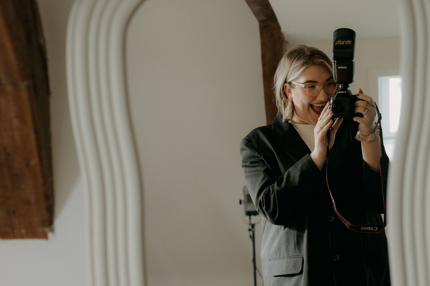 Woman with glasses taking a mirror selfie with a DSLR camera, wearing a dark blazer in a bright room.