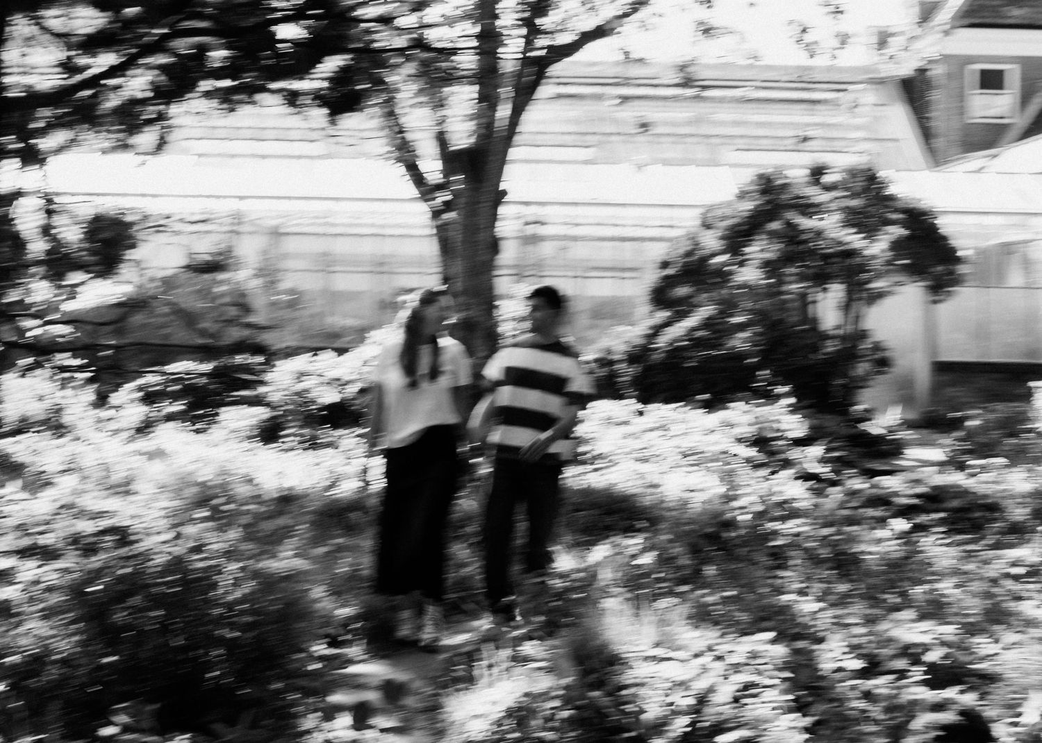 Black and white blurred photo of a couple walking hand in hand among trees and greenery.