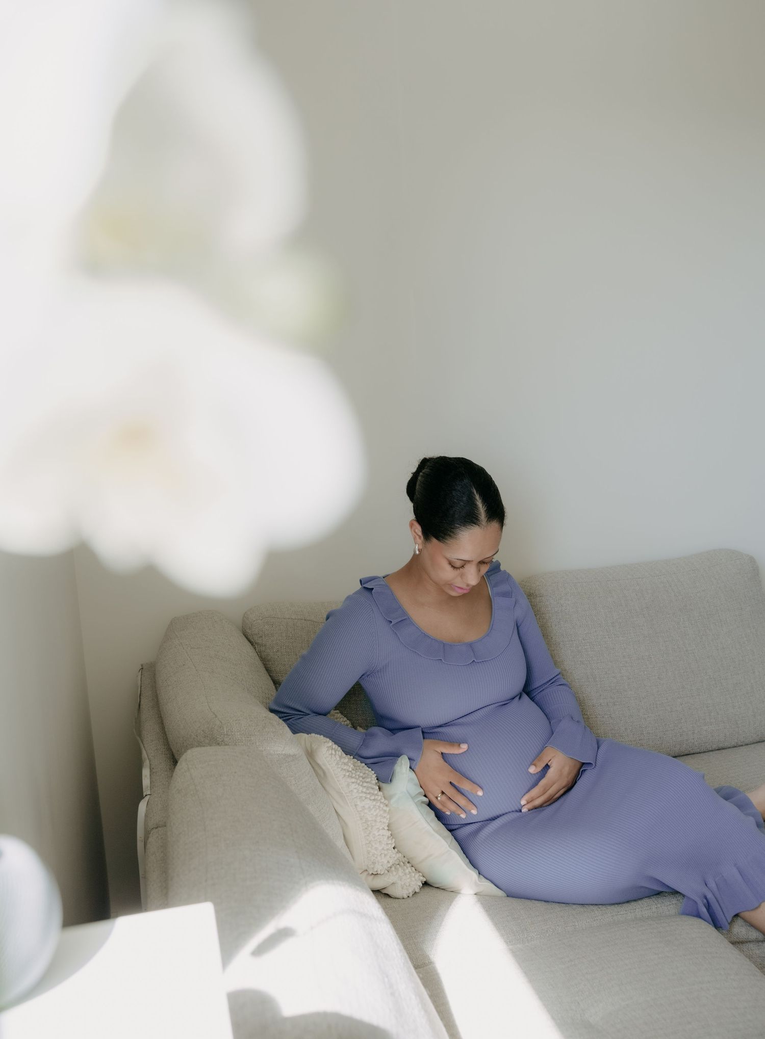 Pregnant woman in lavender dress cradles her baby bump while relaxing on a neutral sofa.