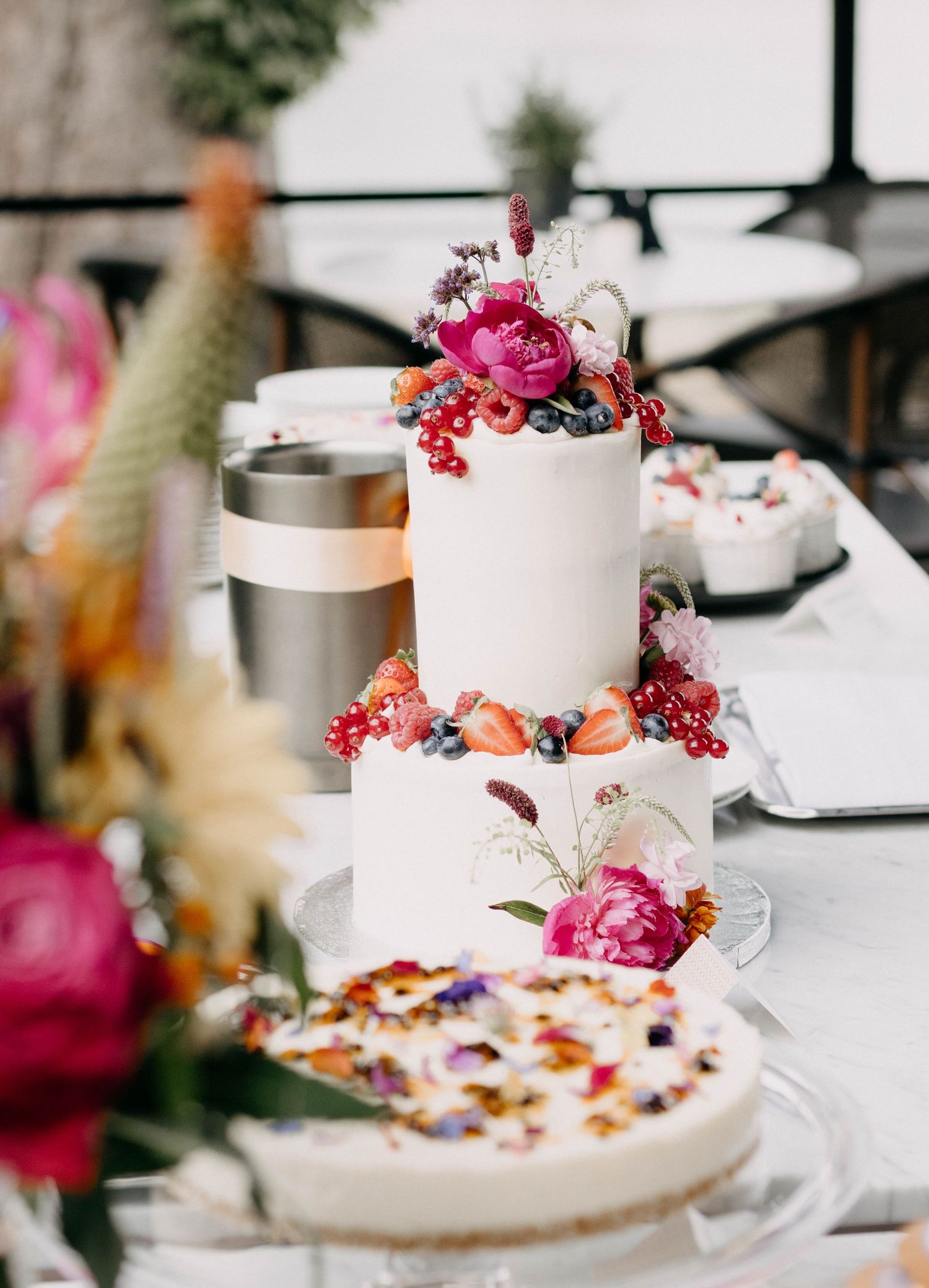 Two-tier white wedding cake adorned with pink peonies, strawberries, and berries on a decorated reception table.