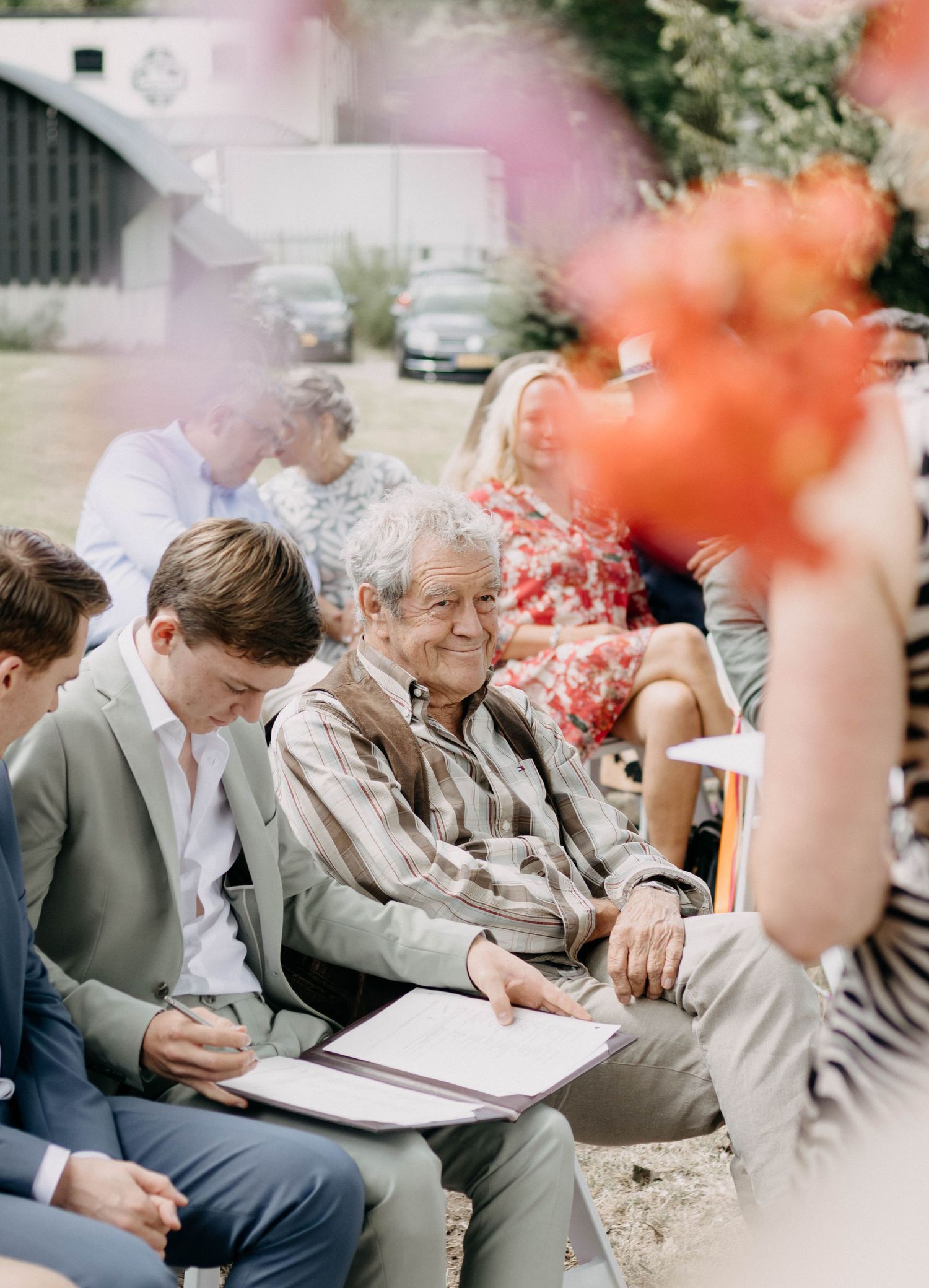 Wedding guests seated outdoors, including an elderly man smiling, with colorful flowers in foreground.