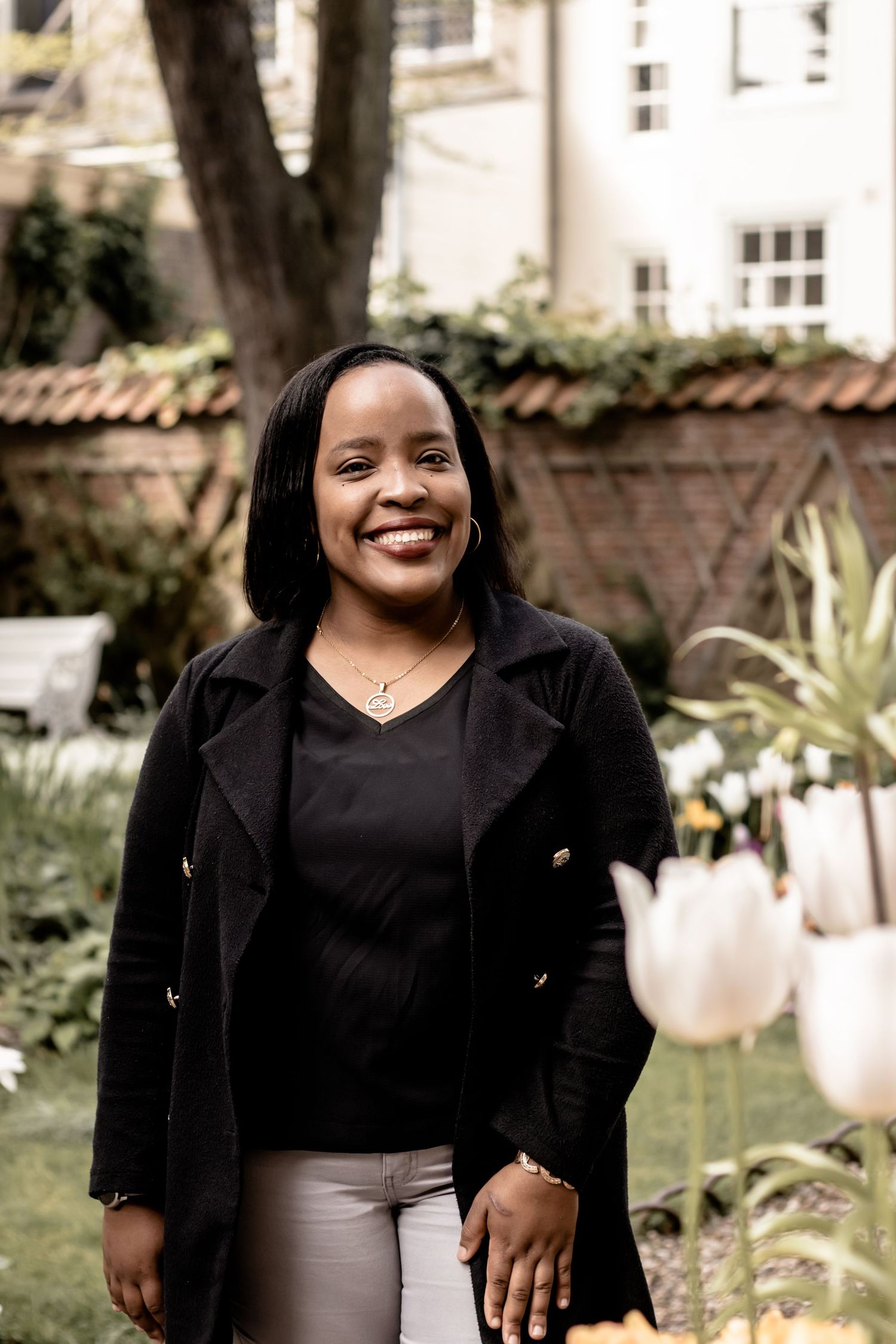 Smiling woman in black coat standing in a garden with tulips and a brick building in the background.