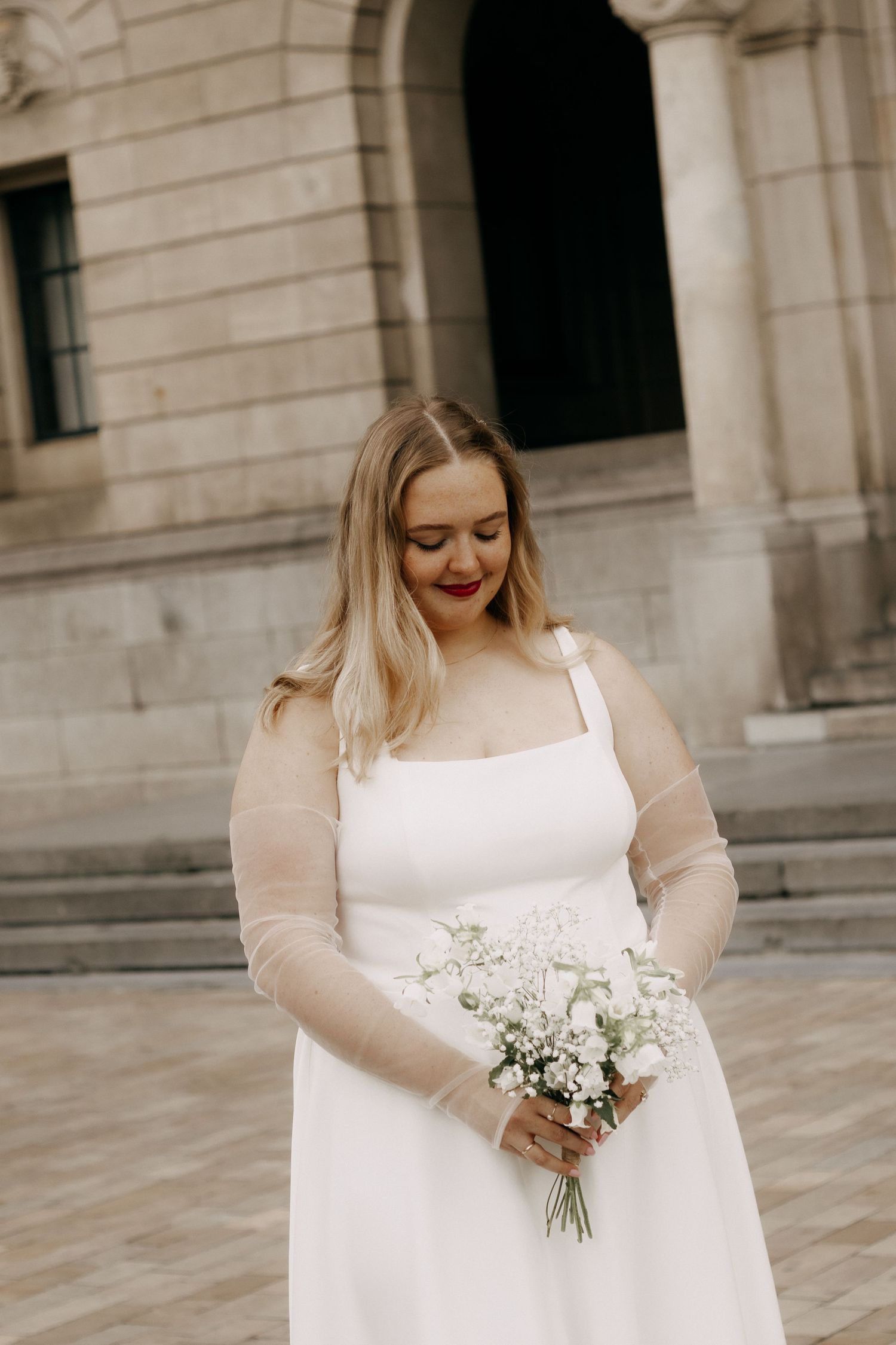 Blonde bride in elegant white gown with sheer sleeves holding a delicate white bouquet on steps.