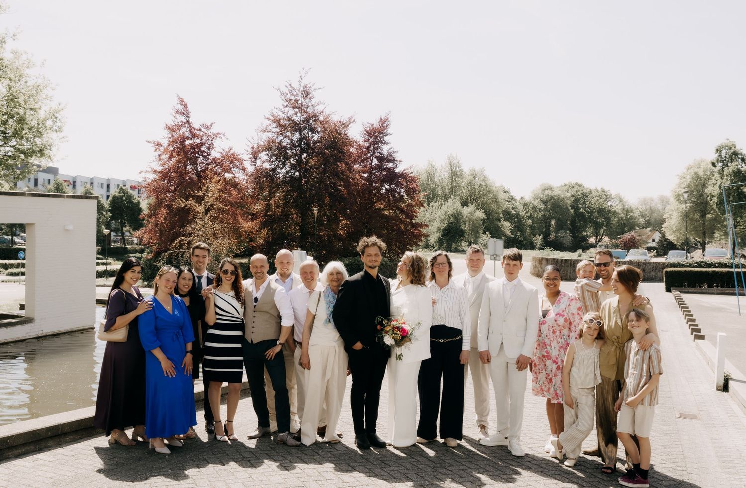 Group wedding photo of couple and guests posing outdoors near a waterway on sunny day.