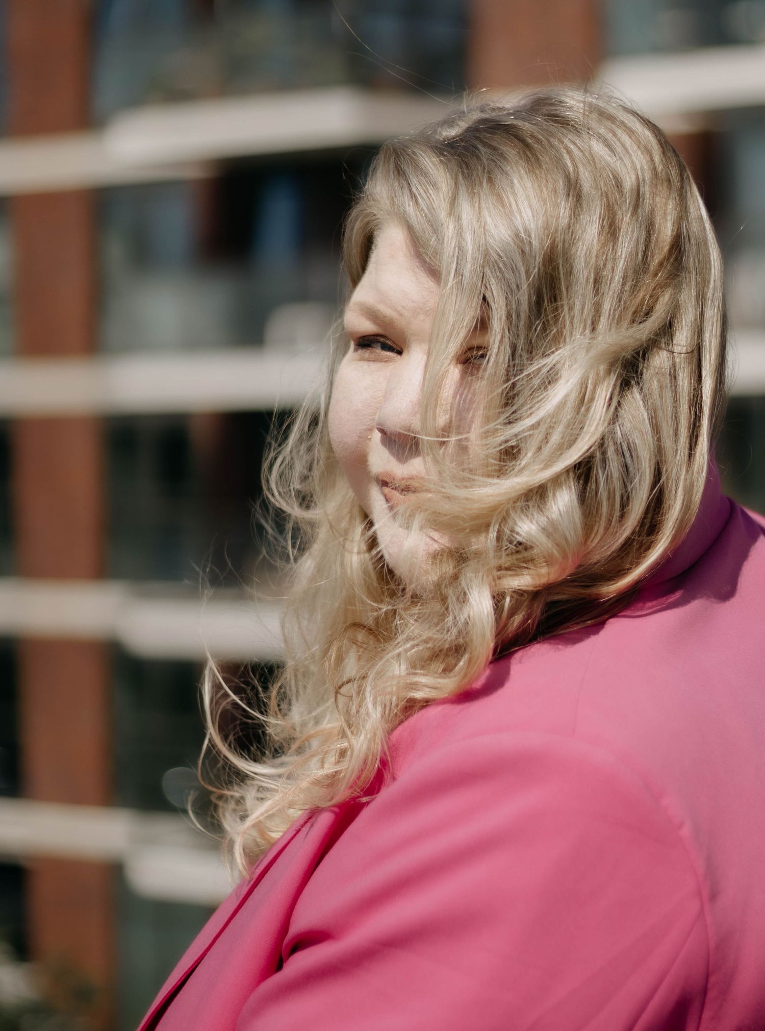 Blonde woman in pink blazer with windswept hair posing outdoors in soft sunlight.