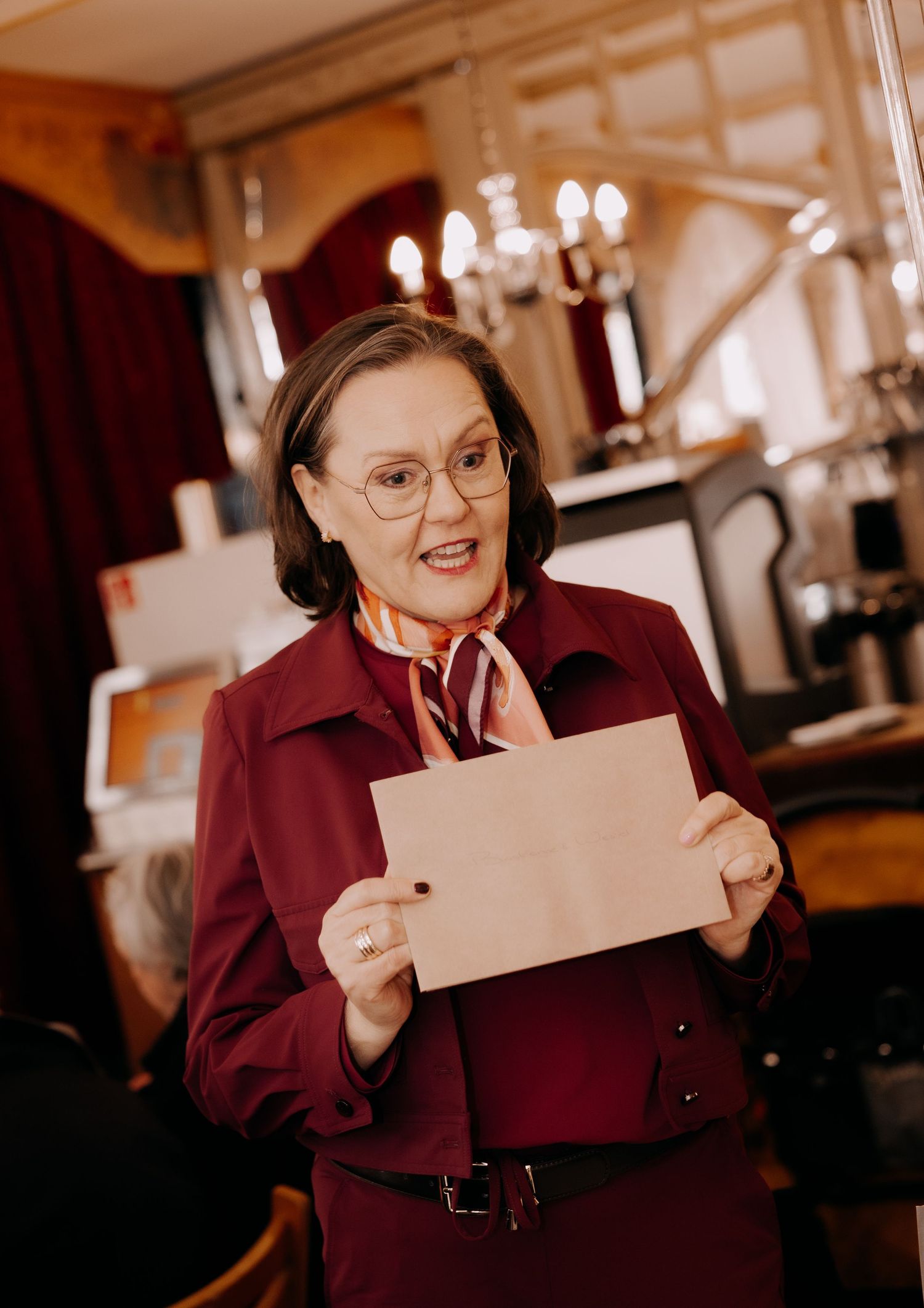 Elegant woman in burgundy suit holding envelope, speaking at indoor wedding reception.