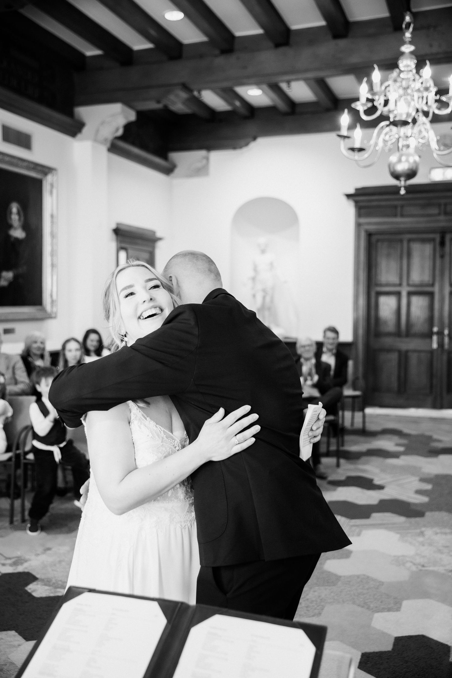 Black and white photo of a joyful bride hugging her groom after their wedding ceremony in an elegant hall.