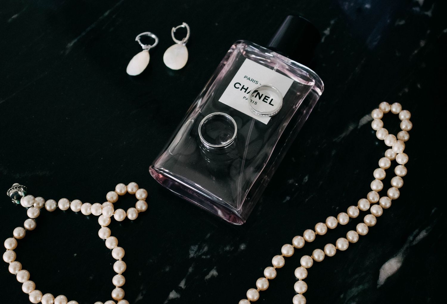 Chanel perfume bottle with pearl necklace and drop earrings on a dark marble surface.