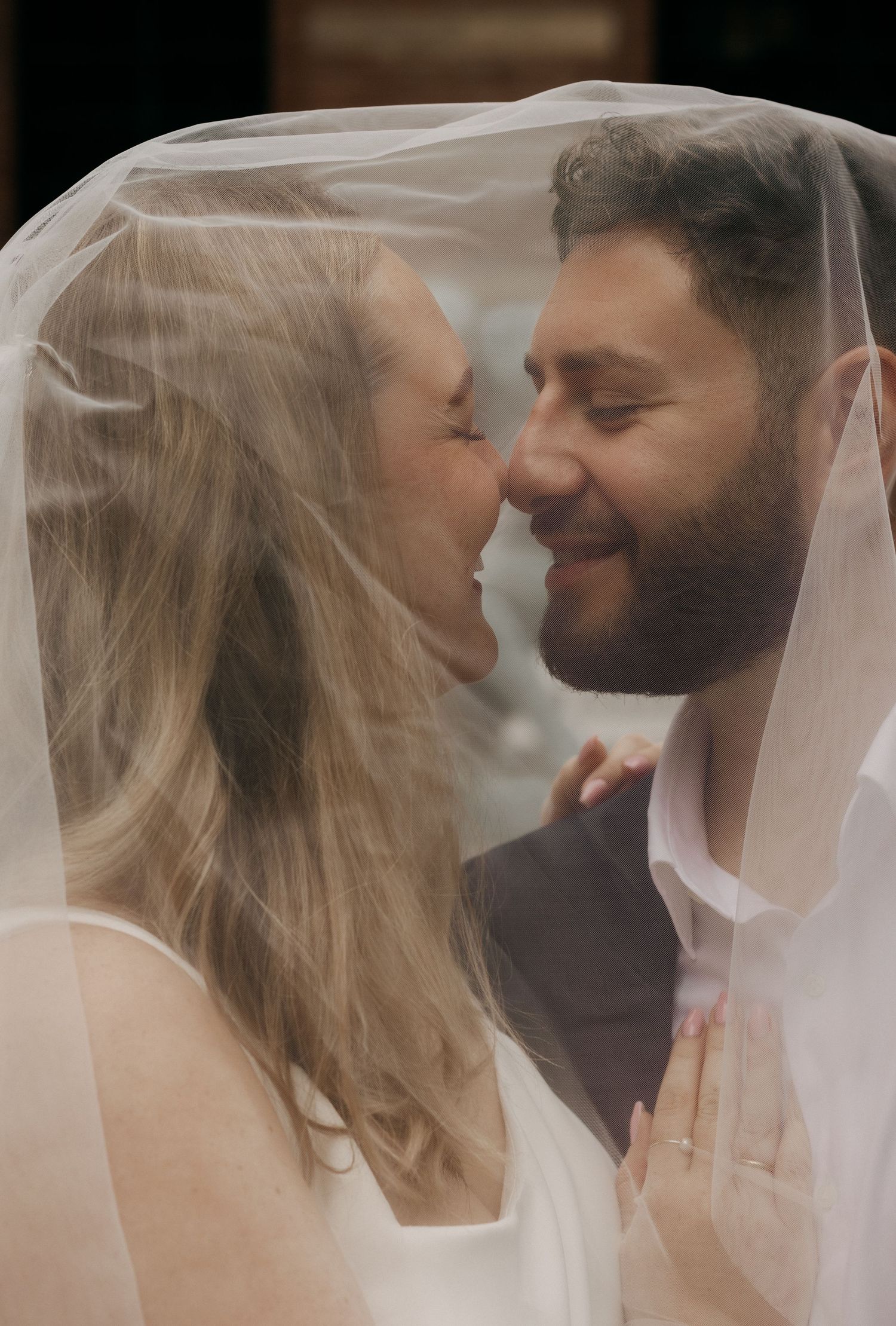 Romantic close-up of bride and groom nearly kissing under sheer wedding veil together.