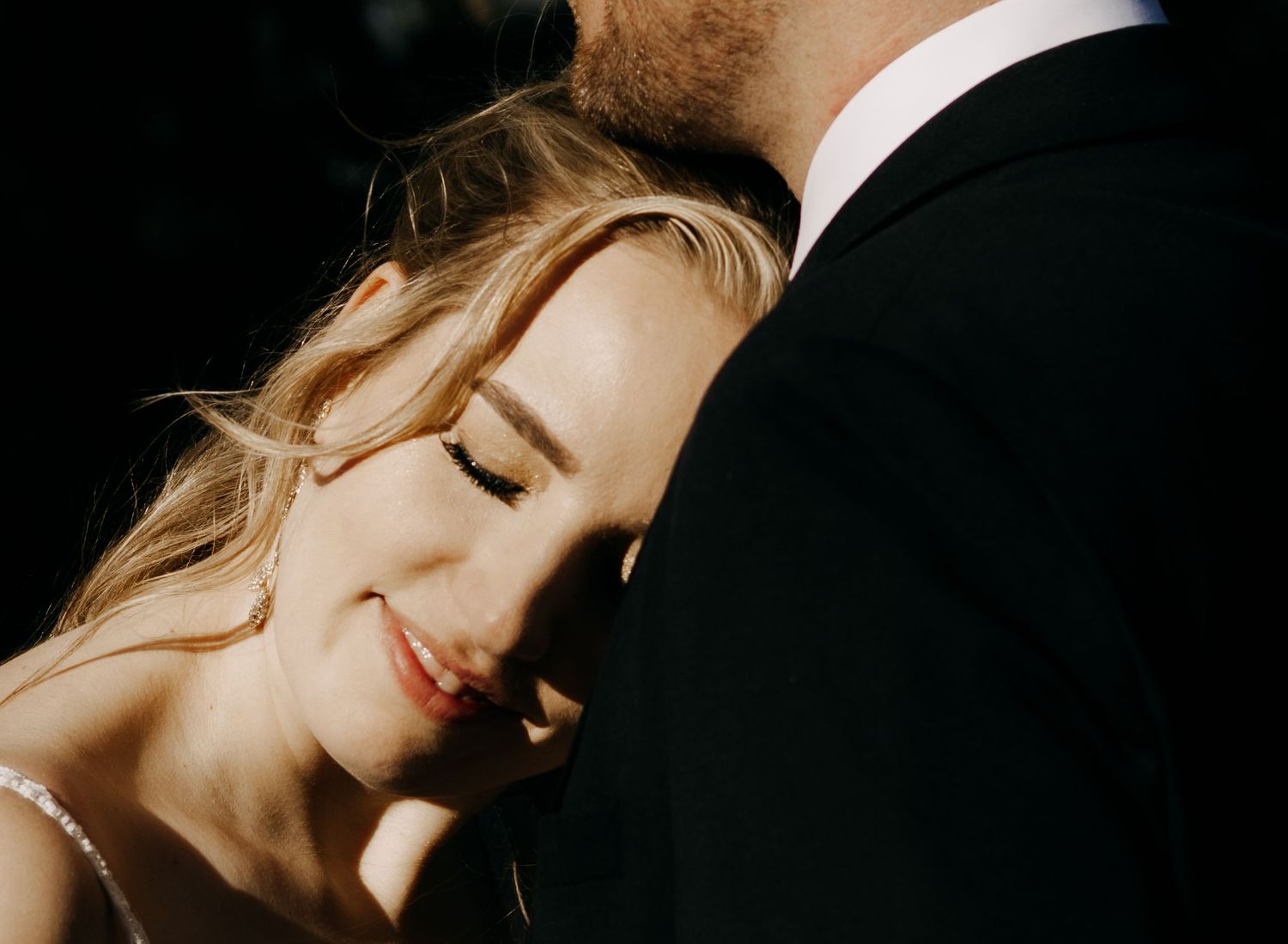 Bride smiles softly while leaning against groom in a warm, intimate close-up wedding portrait.