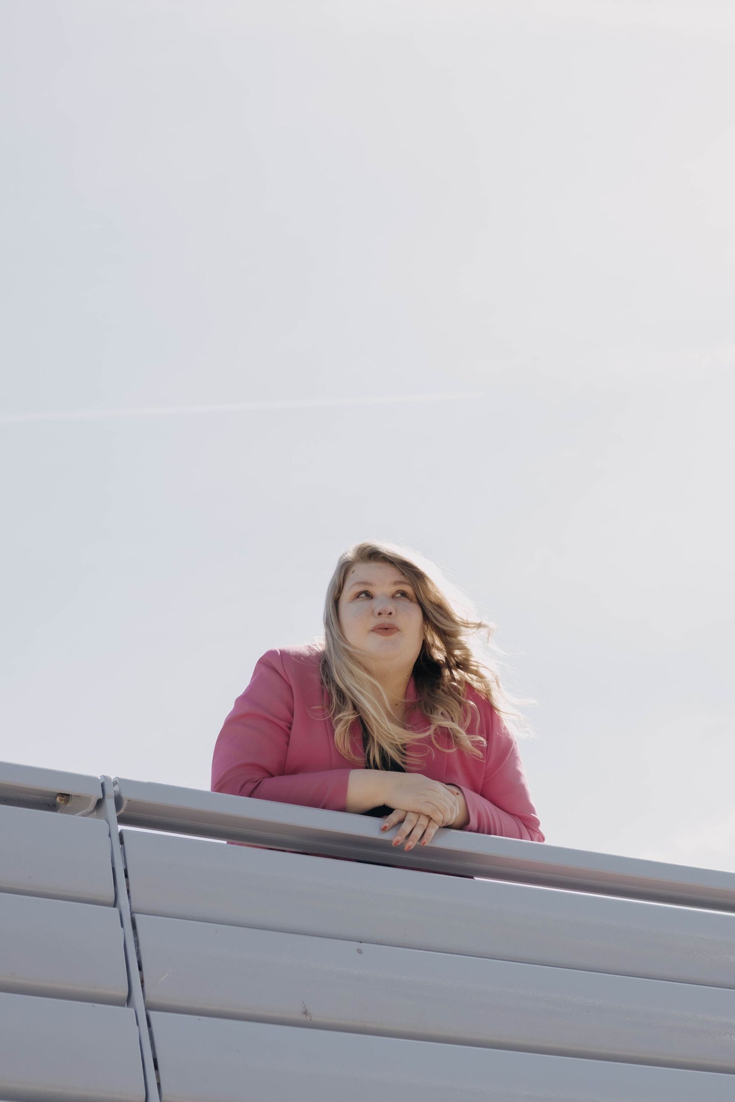 Blonde woman in pink jacket leaning on a railing looking up at a bright sky.