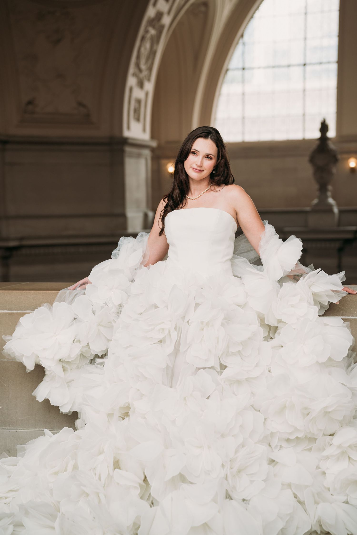 Bride in a voluminous white ruffled ball gown poses gracefully on marble steps in an elegant grand hall.