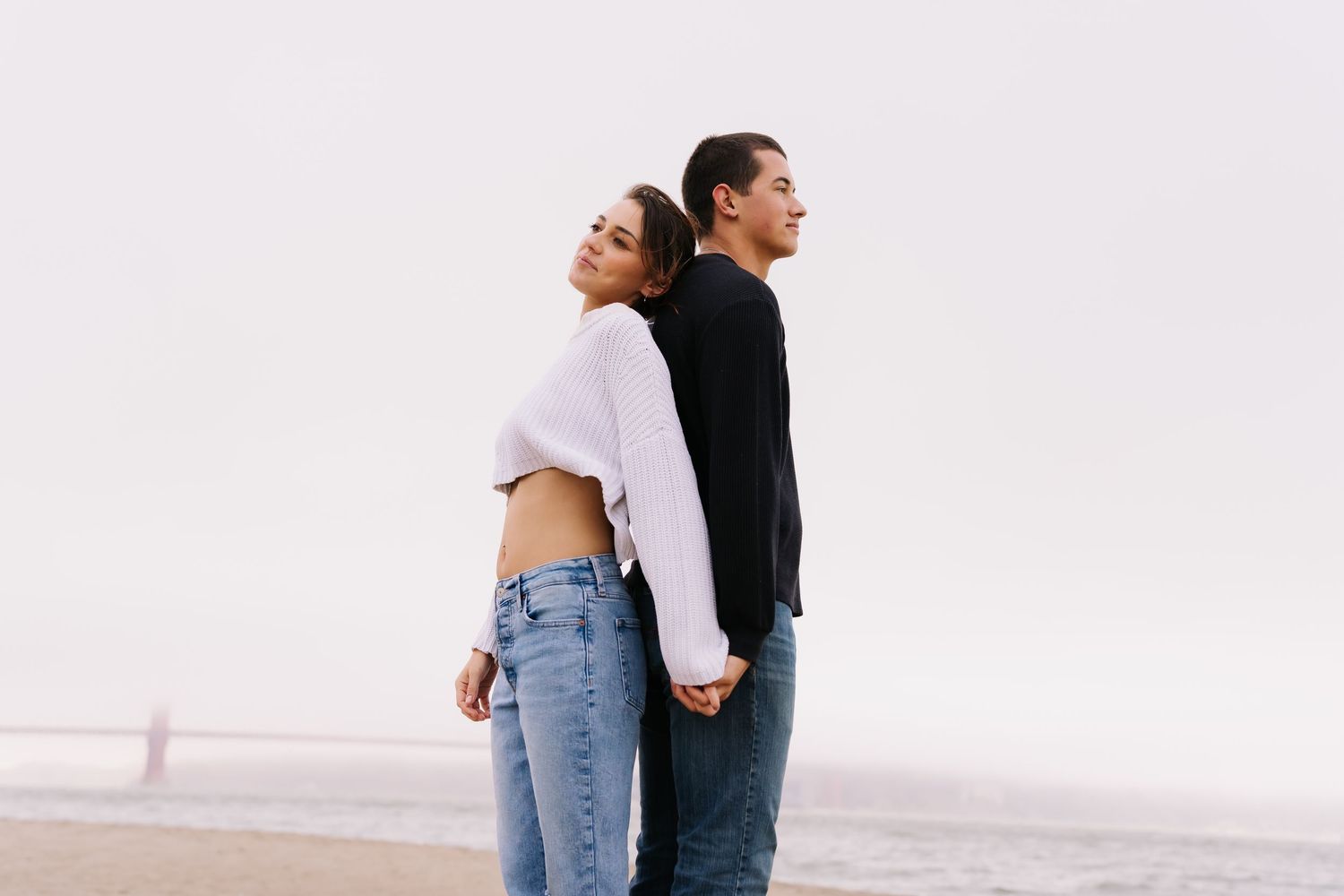 Couple standing back-to-back on a beach, holding hands, wearing casual jeans and tops on an overcast day.