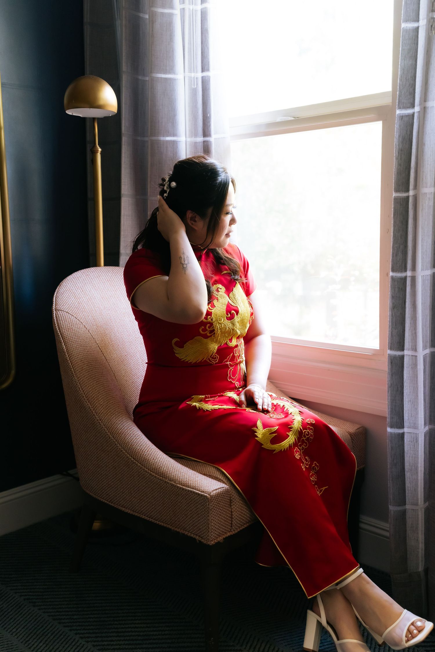 Woman in red Chinese qipao dress with gold dragon embroidery sits by window in pink chair, touching her hair.