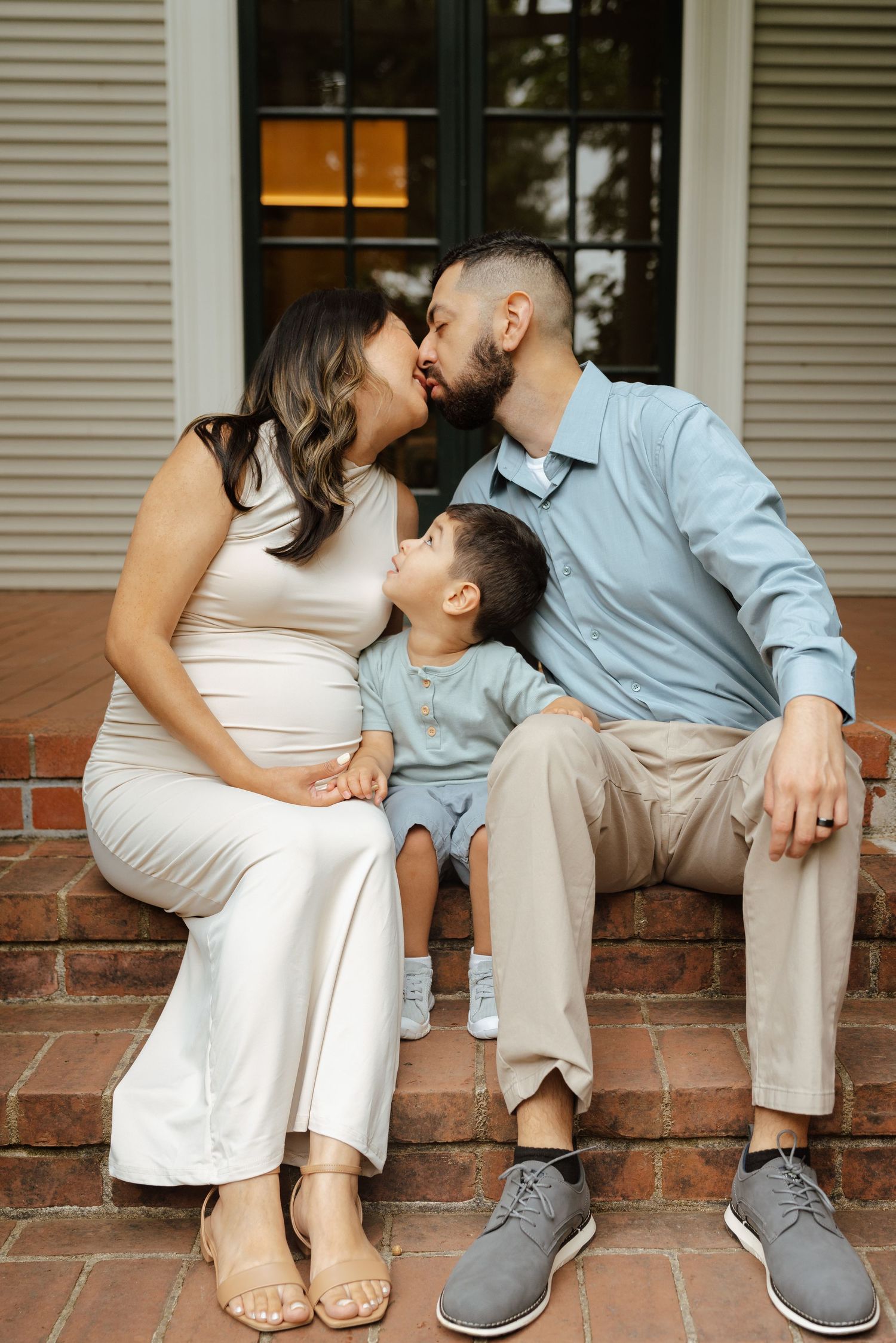 Pregnant woman and man kissing on brick steps while young toddler boy looks up at them during family maternity photo session.