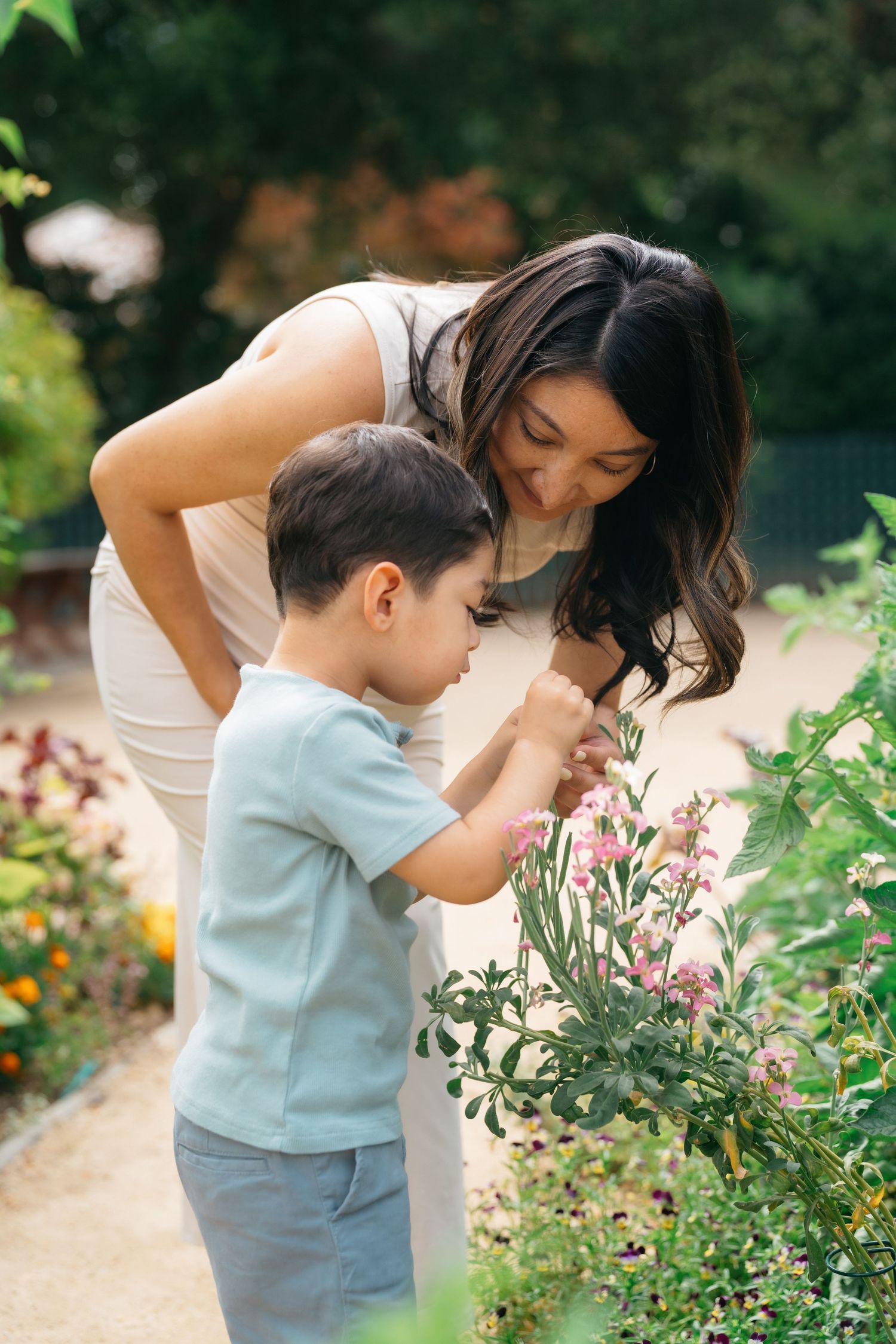 A mother and young son explore a garden together, smelling pink flowers among lush green plants on a sunny day.