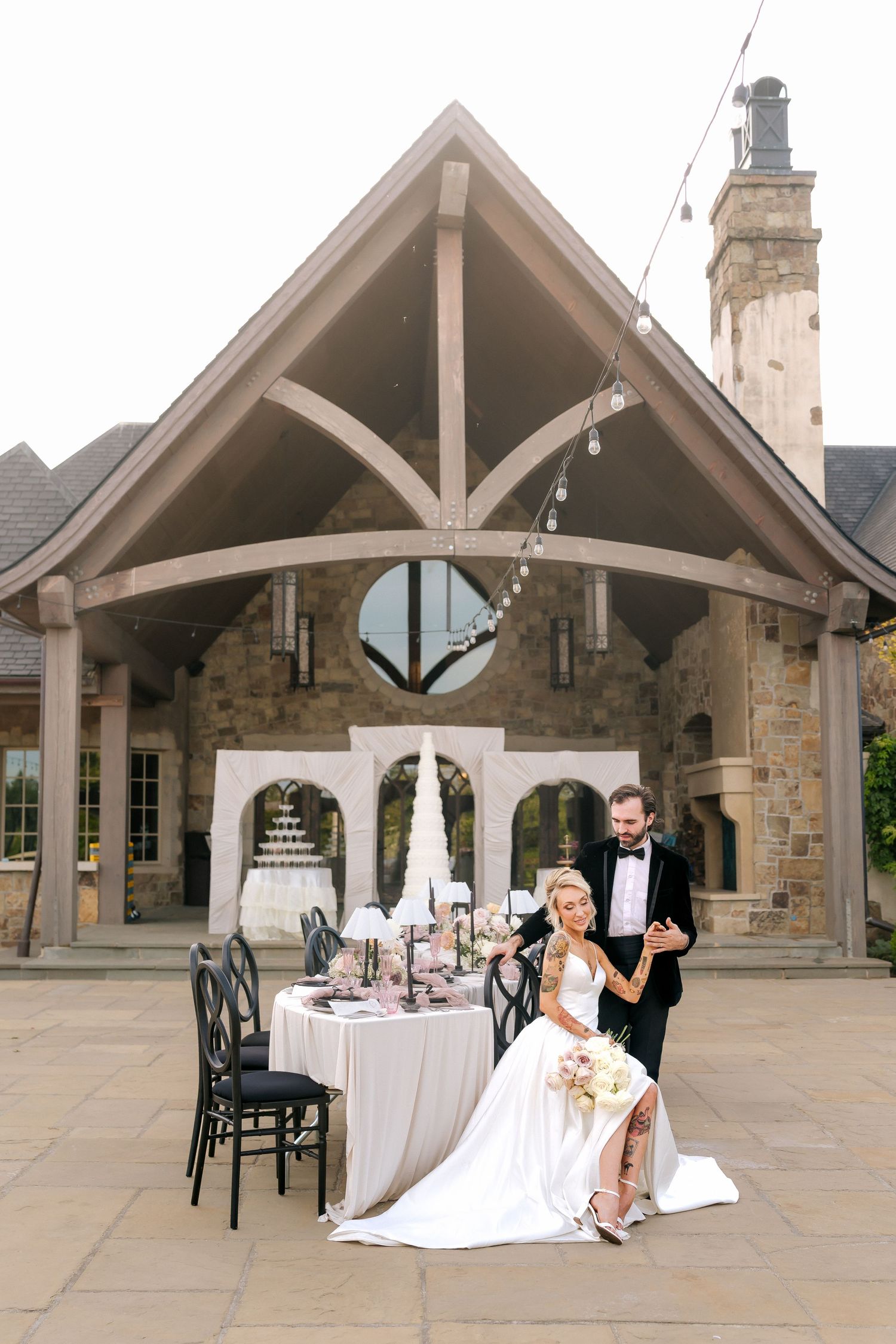Bride and groom pose at elegant outdoor sweetheart table with stone chapel backdrop and string lights at luxury wedding venue.