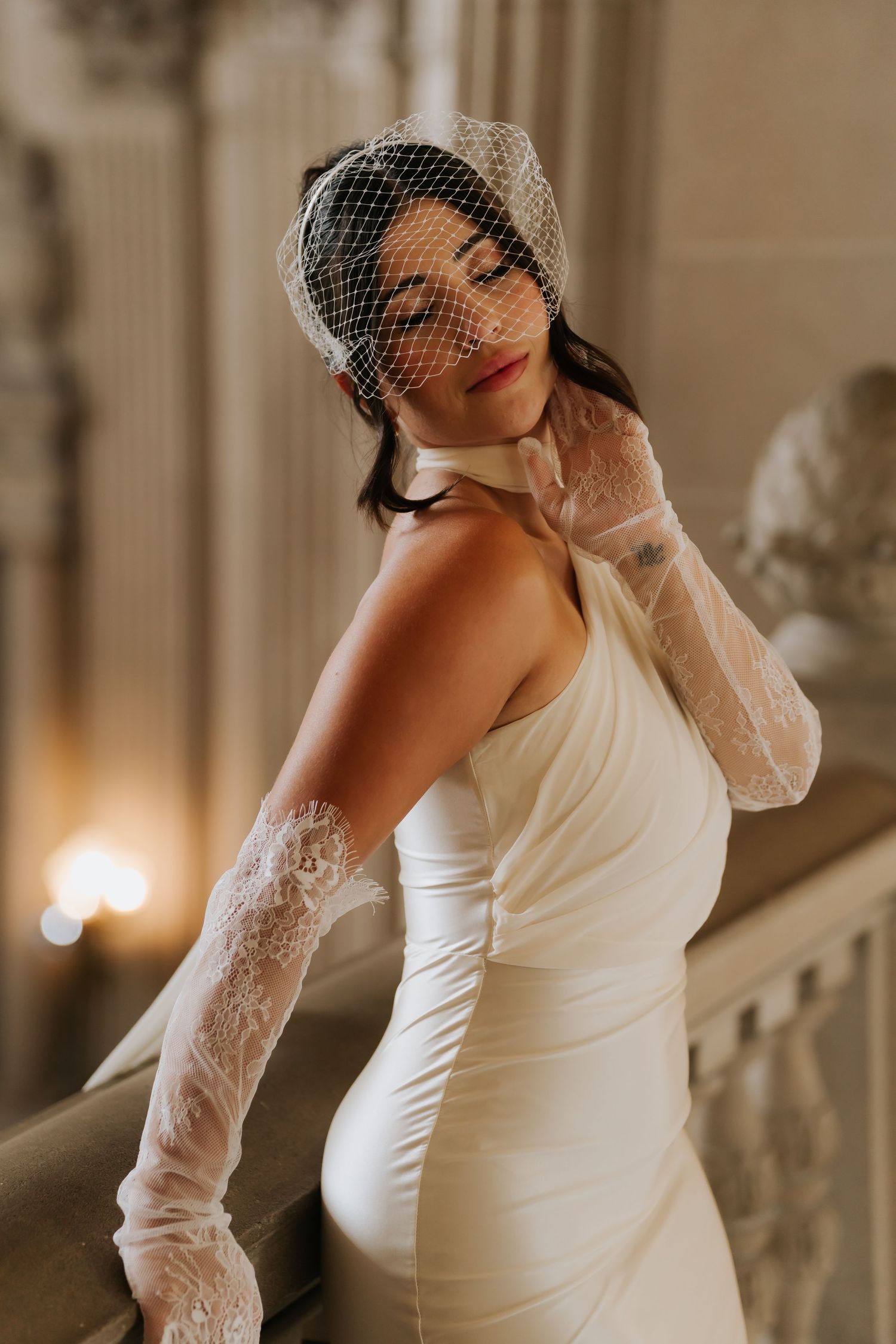 Bride in white satin gown, lace gloves, and birdcage veil poses elegantly on ornate staircase.