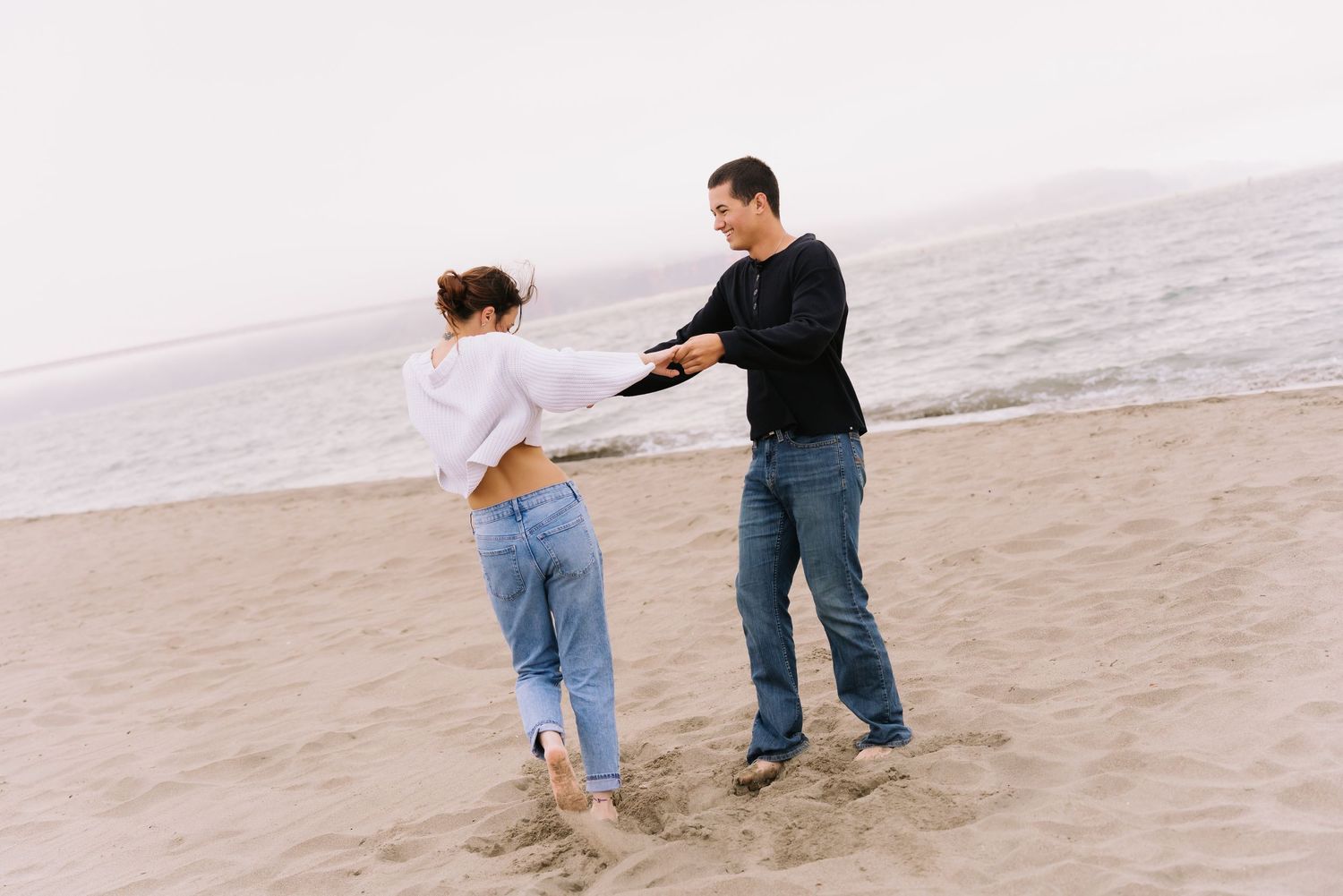 Couple dancing playfully on a sandy beach with ocean waves in the background on an overcast day.