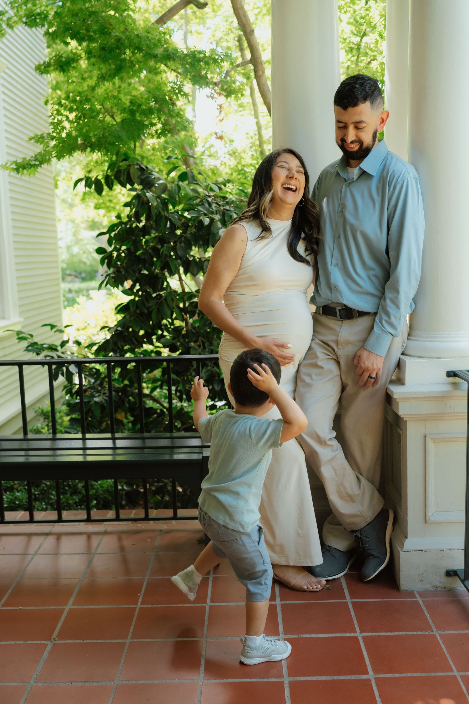 Pregnant woman laughing with husband and toddler child on a porch surrounded by green trees.