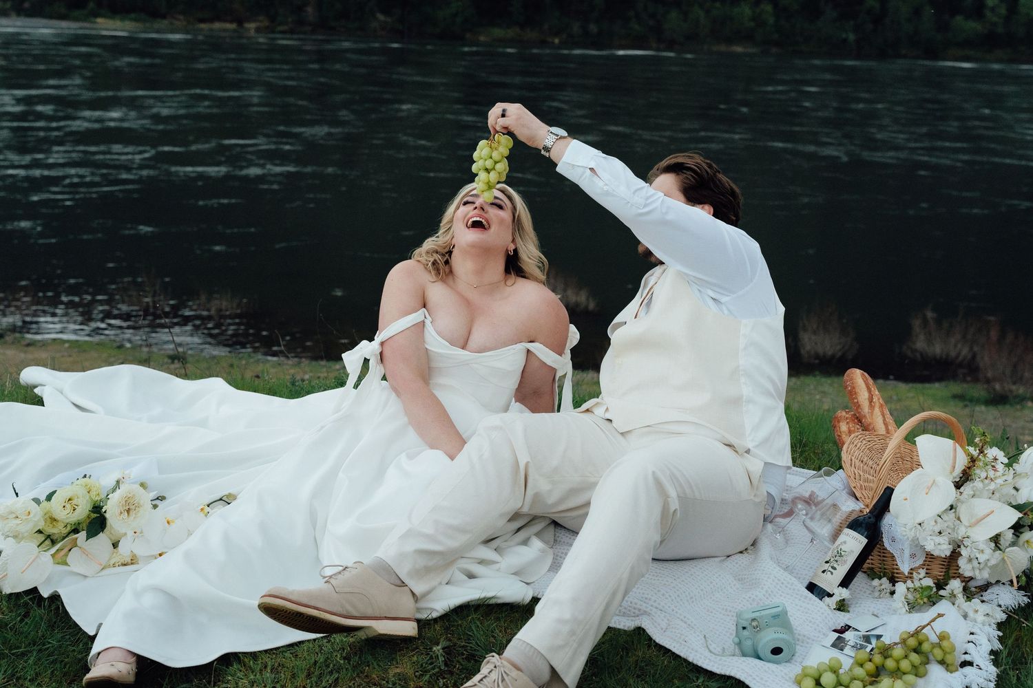 Joyful bride laughs as groom feeds her grapes during romantic riverside picnic.