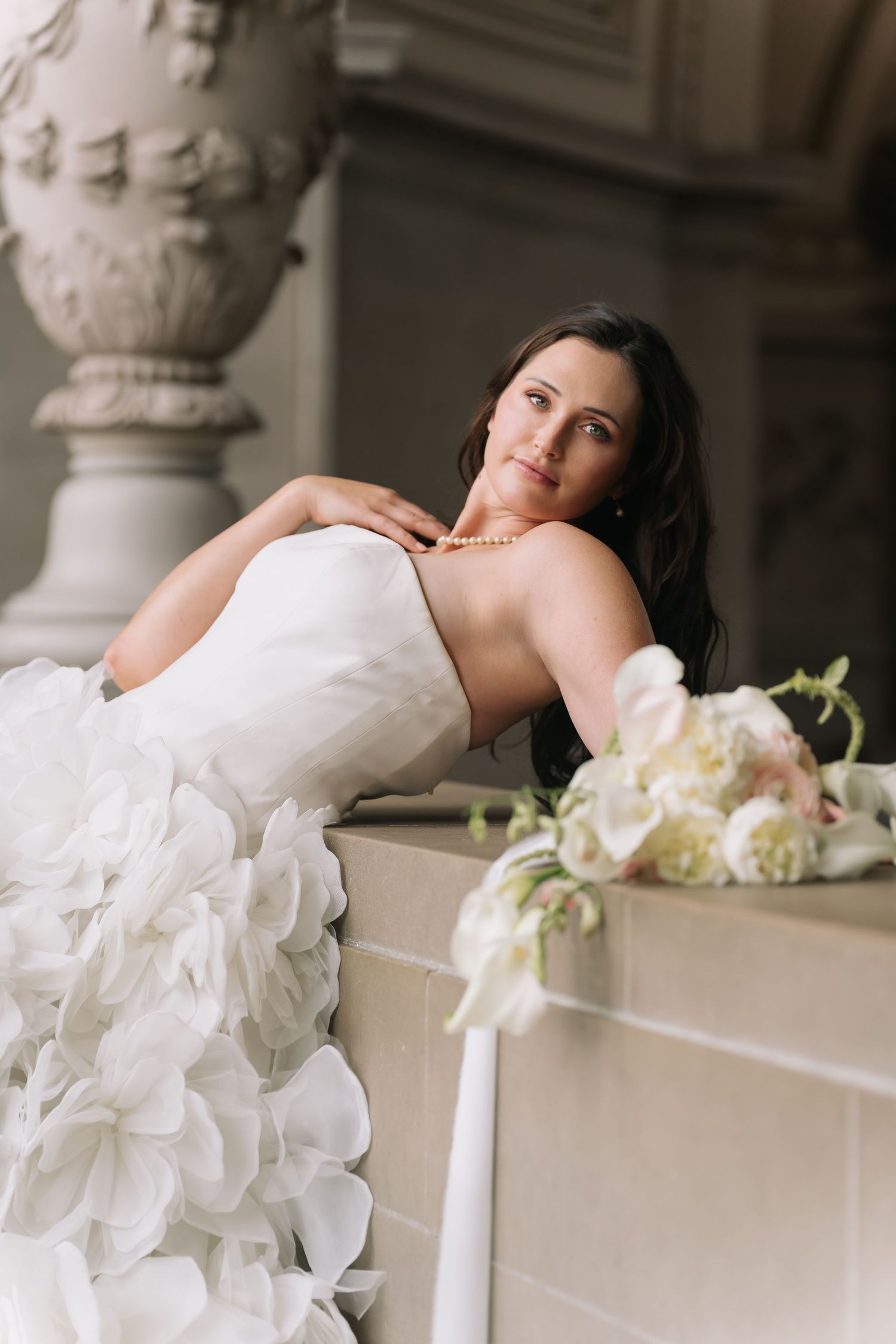 Elegant bride in strapless white ruffled gown with pearl necklace, leaning gracefully near floral bouquet in ornate venue.