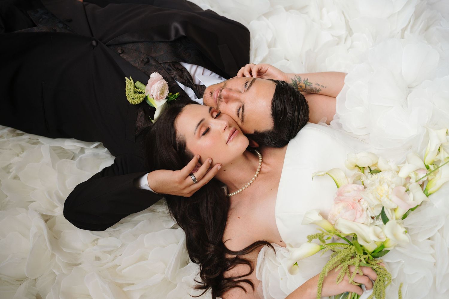Bride and groom lying on white wedding dress, sharing a tender moment with pink flowers.