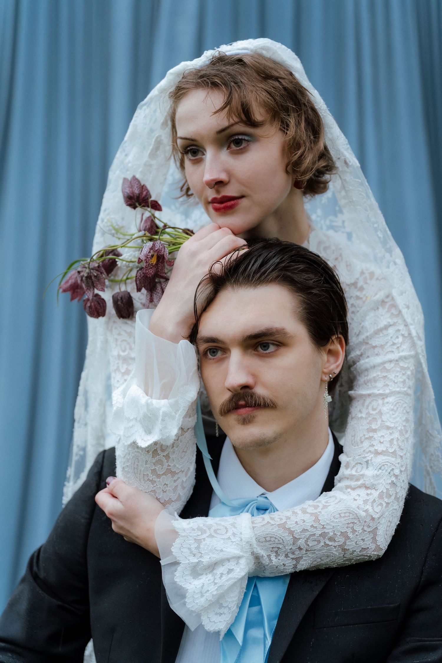Vintage wedding portrait of bride in lace veil holding dark flowers, embracing groom with mustache in black suit.