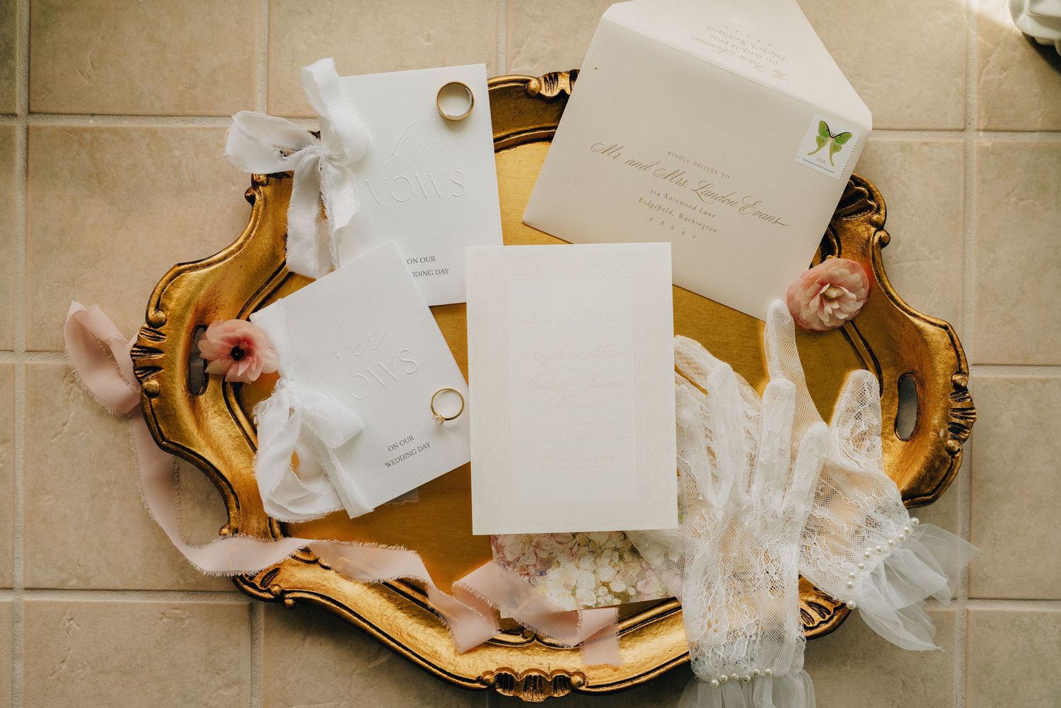 Elegant wedding stationery flatlay with white envelopes, gold rings, pink flowers, and ribbons on ornate gold tray.