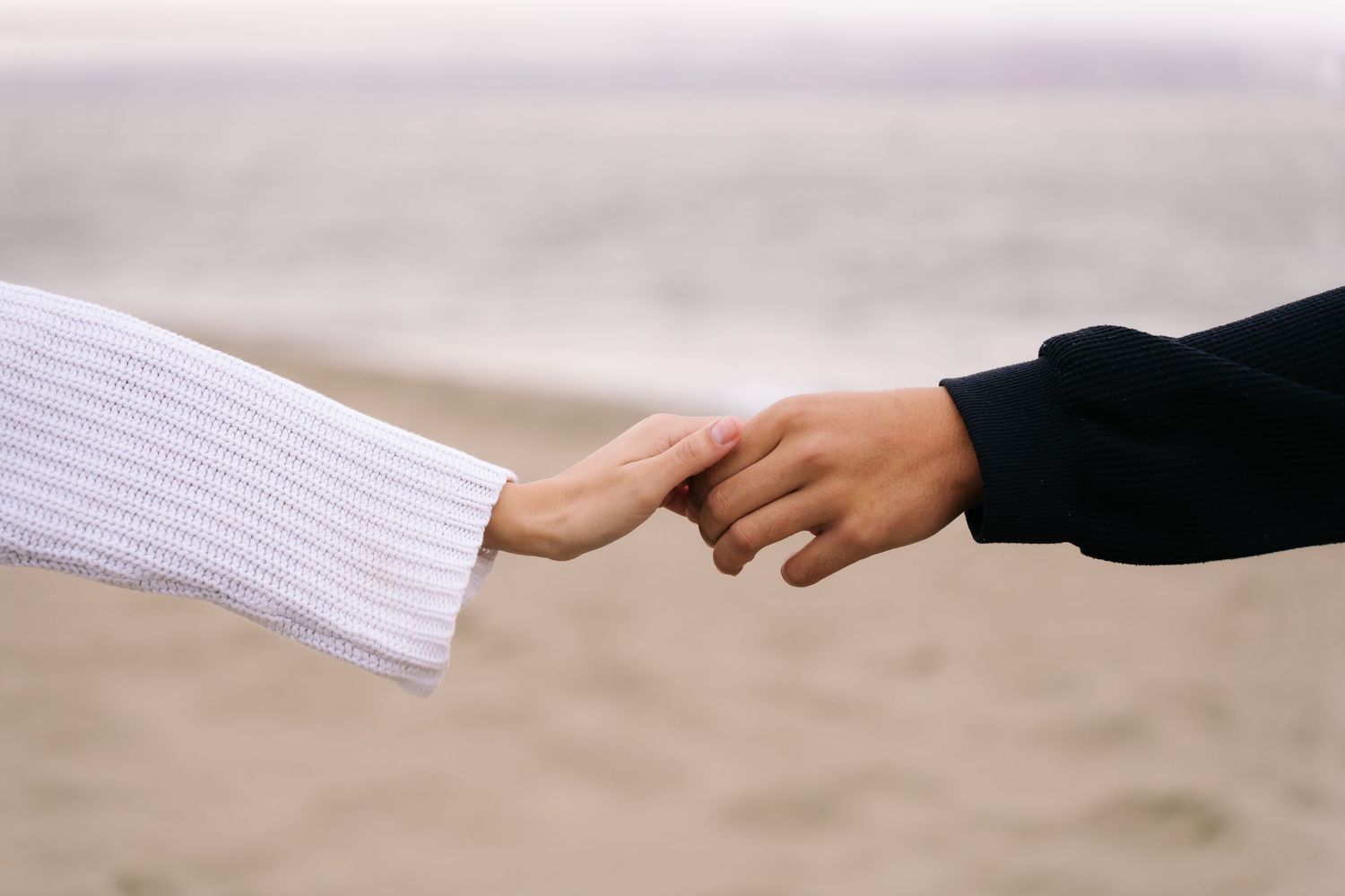 Two people gently holding hands on a sandy beach, one wearing a white knit sweater and the other a dark navy top.