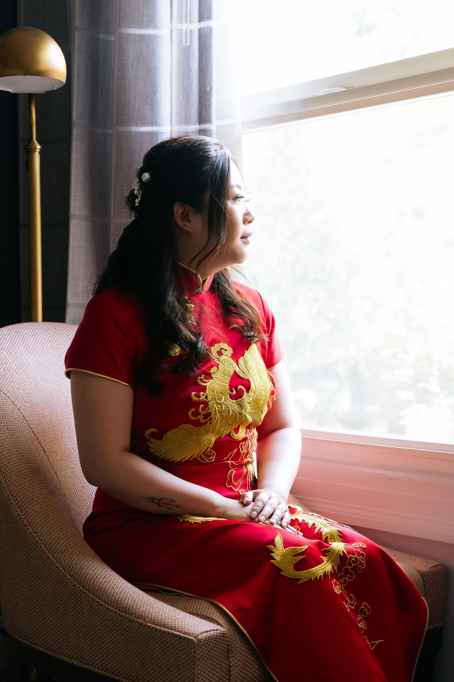 Woman in red embroidered Chinese qipao dress sitting by window in soft natural light.
