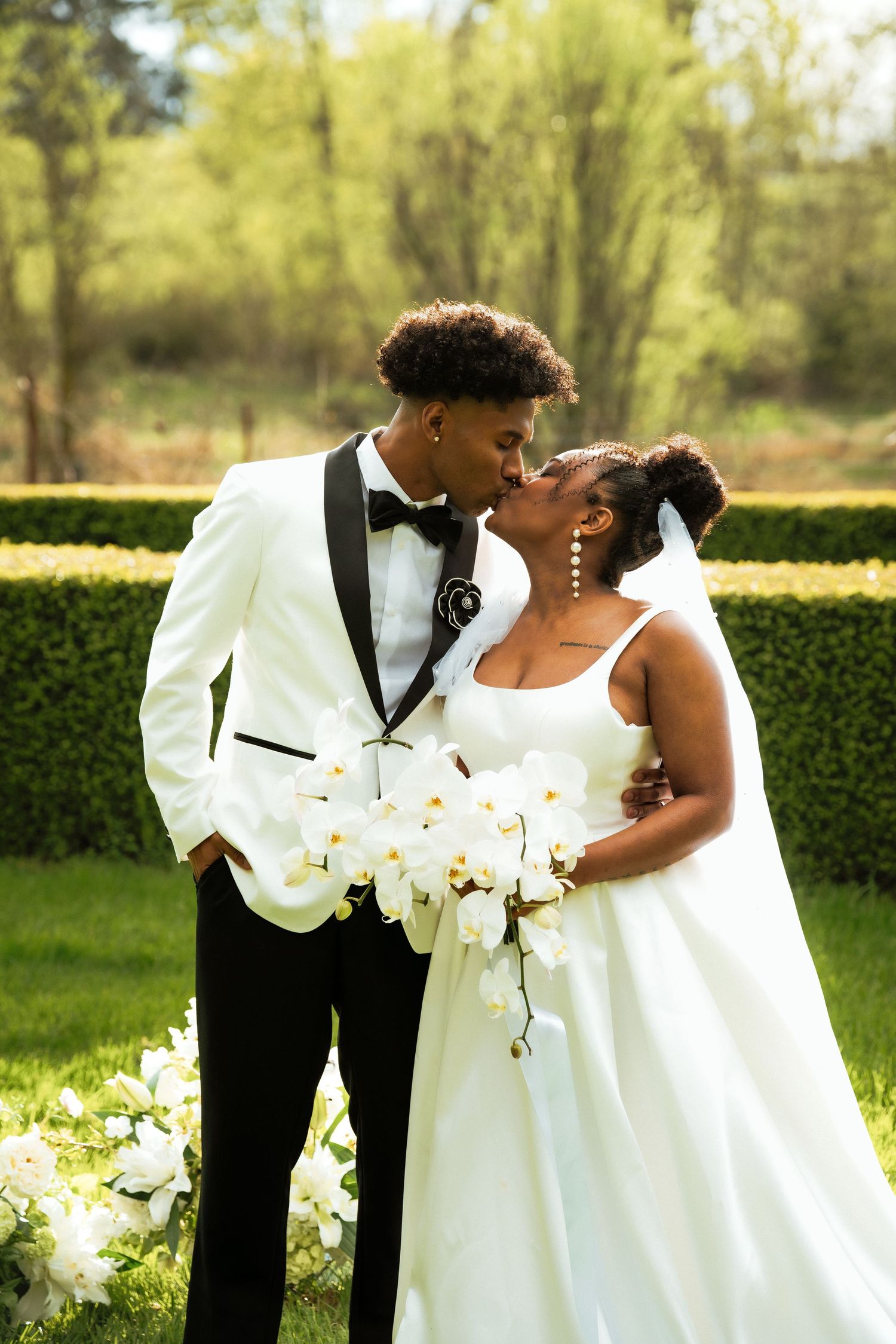 Black couple shares kiss in lush green garden, bride holds white orchid bouquet.