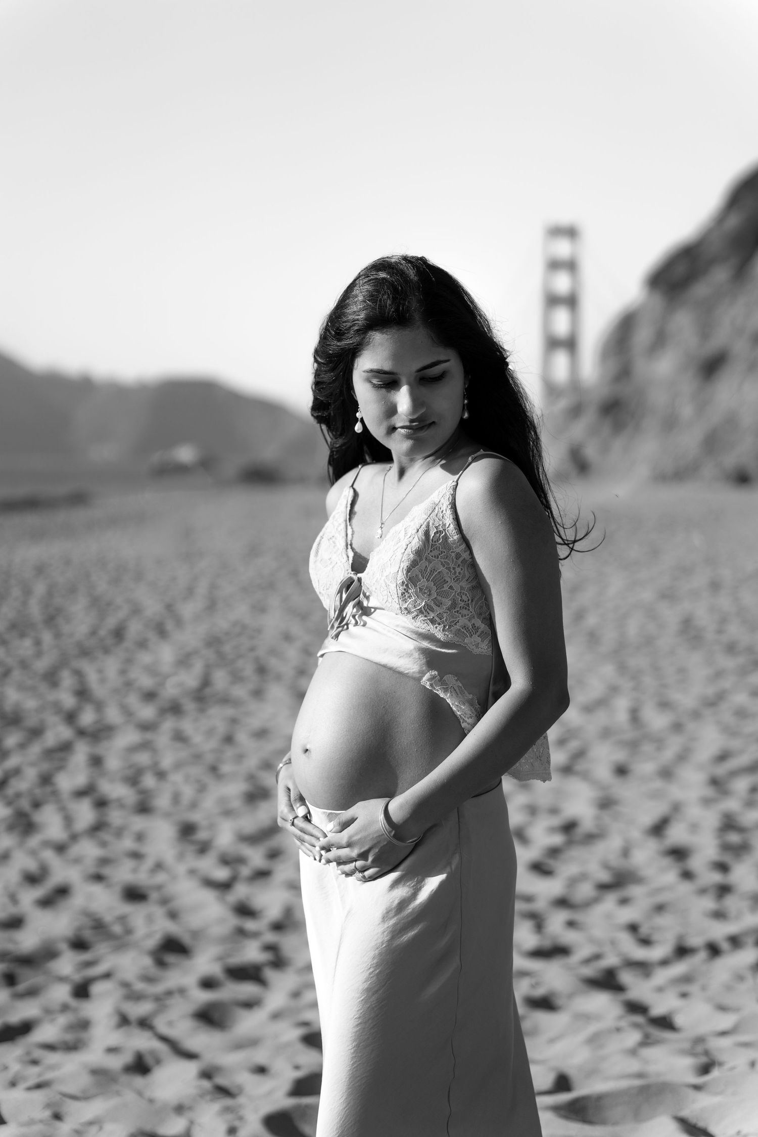 Black and white photo of pregnant woman cradling belly on sandy beach near Golden Gate Bridge in San Francisco.