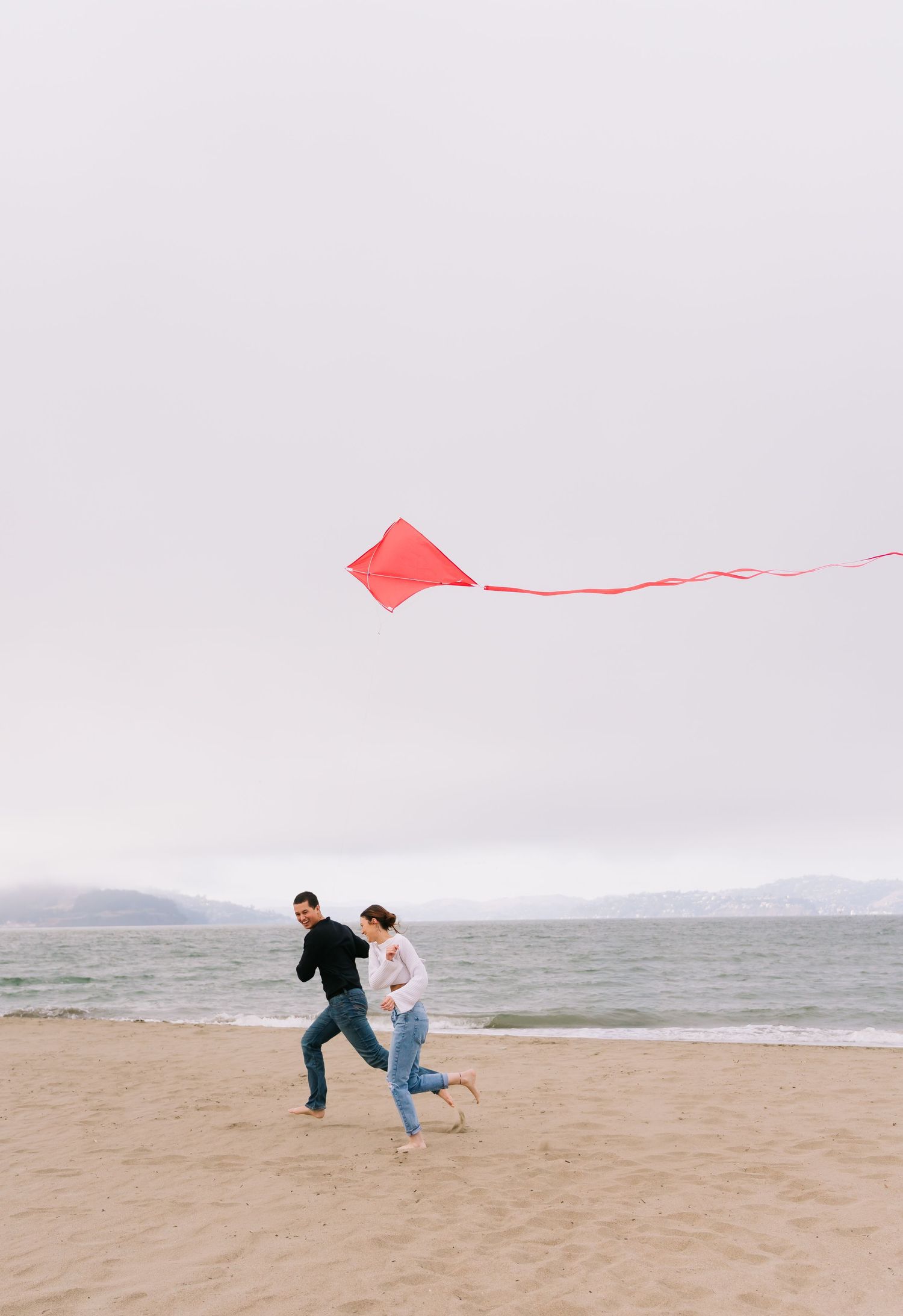 Couple running on a sandy beach flying a red kite on an overcast day with ocean views.