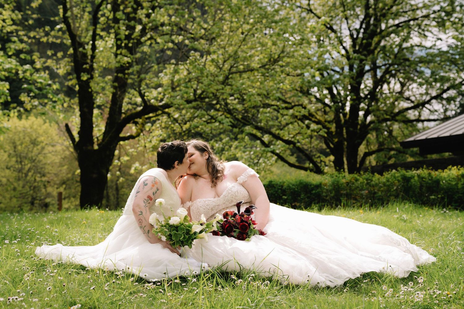 Two brides in white gowns kissing while seated on green grass beneath blooming trees with bouquets.