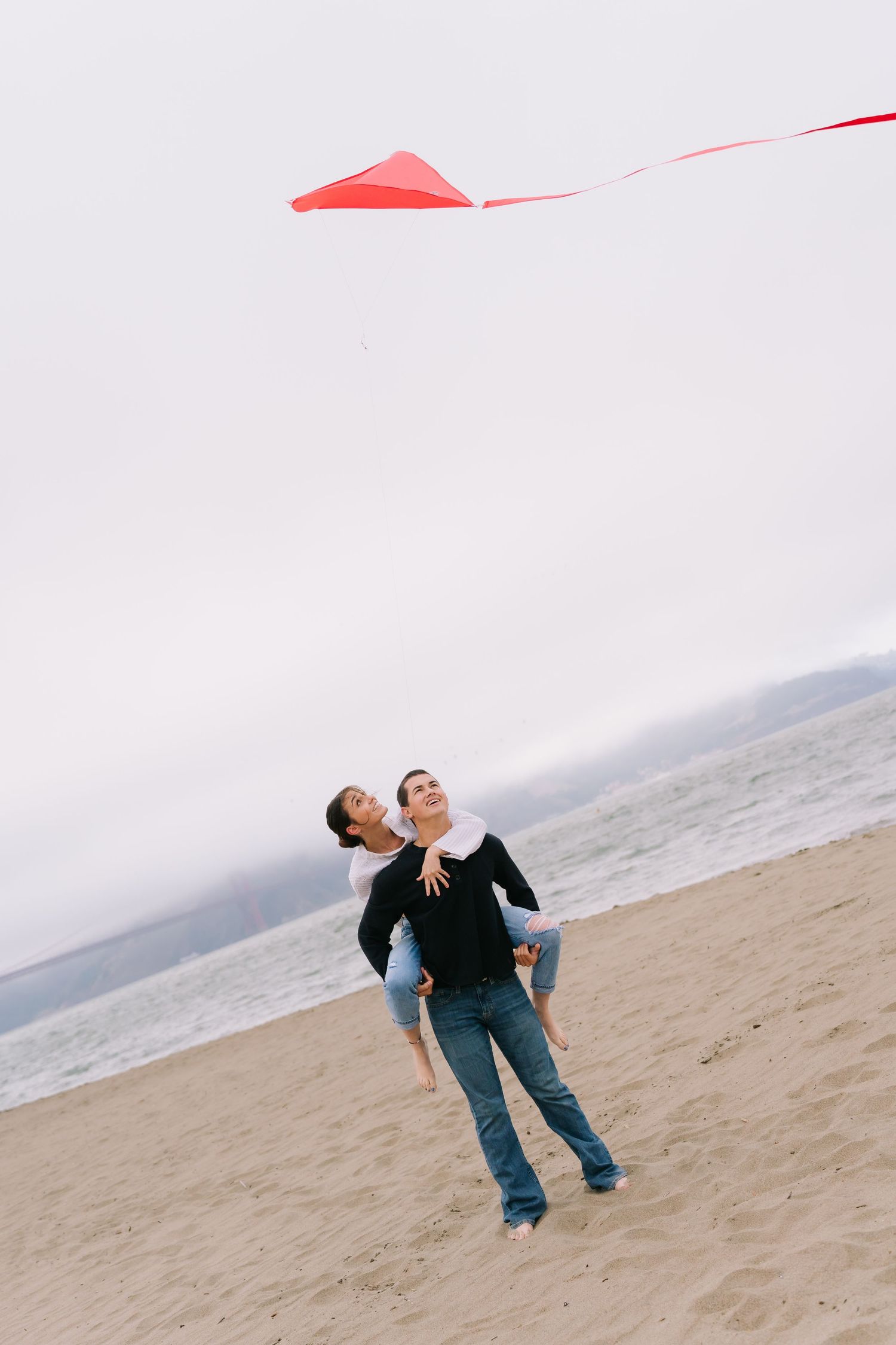 Couple flying a red kite on a sandy beach, laughing and embracing with the ocean in the background.