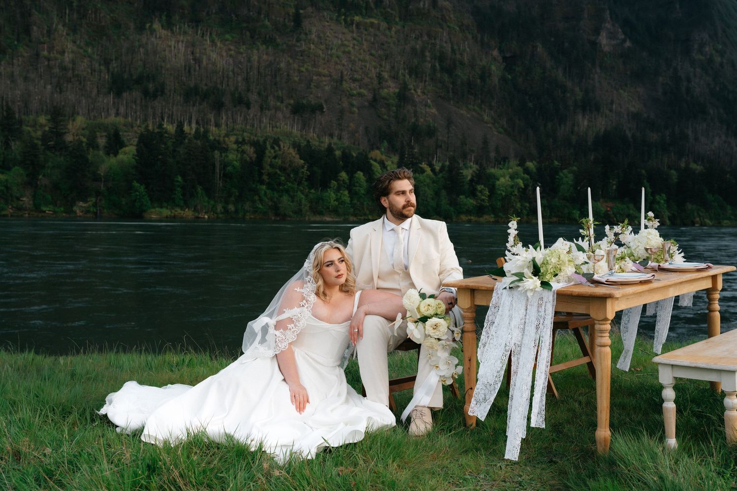 Bride in white gown sits on grass beside groom in ivory suit at rustic table with floral decor near a mountain lake.