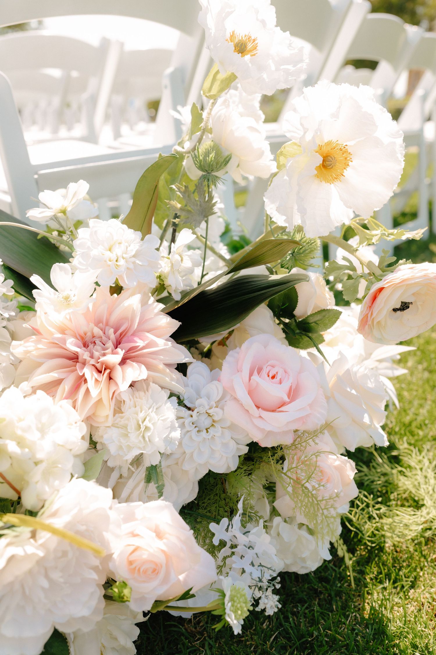 Elegant wedding floral arrangement featuring blush pink roses, white dahlias, and peonies on green grass beside white chairs.