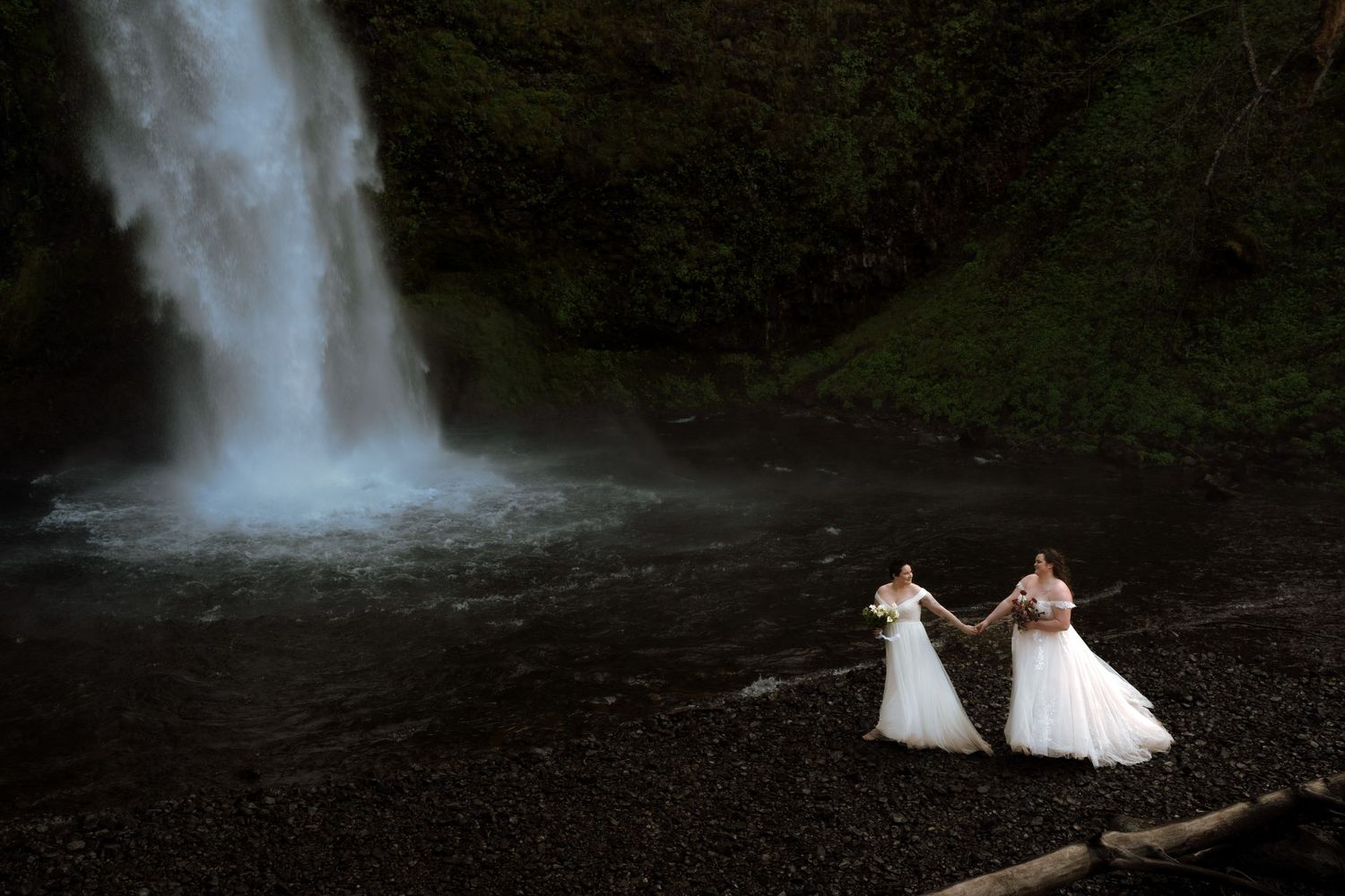 Two brides in white wedding gowns holding hands in front of a dramatic waterfall on rocky ground.
