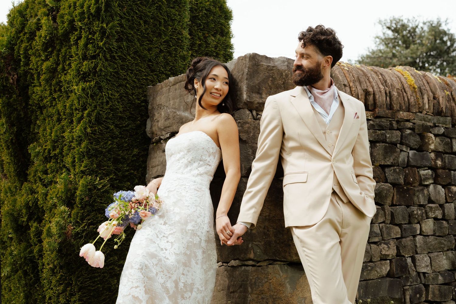 Bride in strapless lace gown and groom in tan suit hold hands near stone wall and hedges.