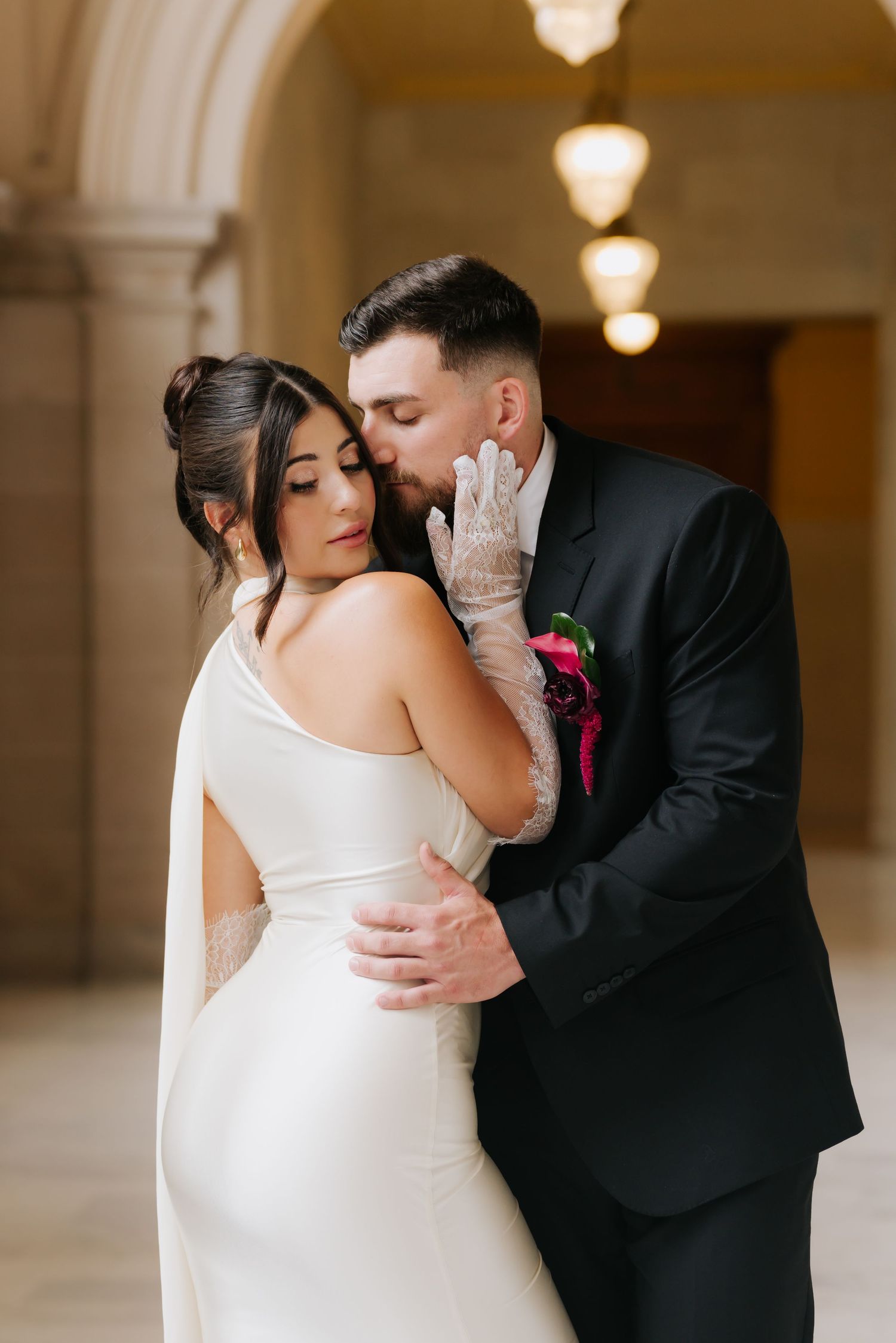 A groom in a black suit kisses his bride in a white gown and lace gloves inside an elegant venue.