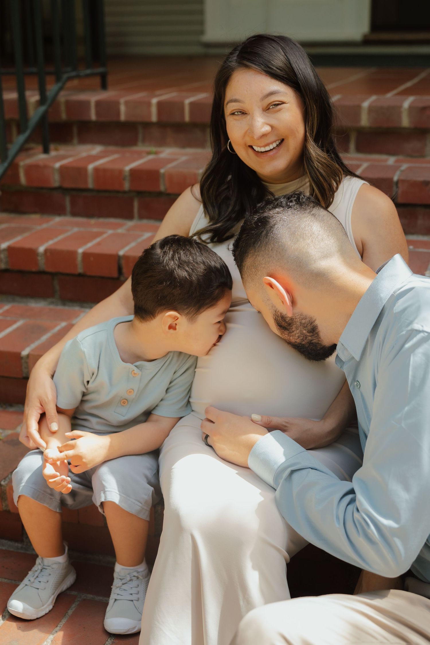 Pregnant woman smiling on brick steps with husband and young son leaning against her baby bump.