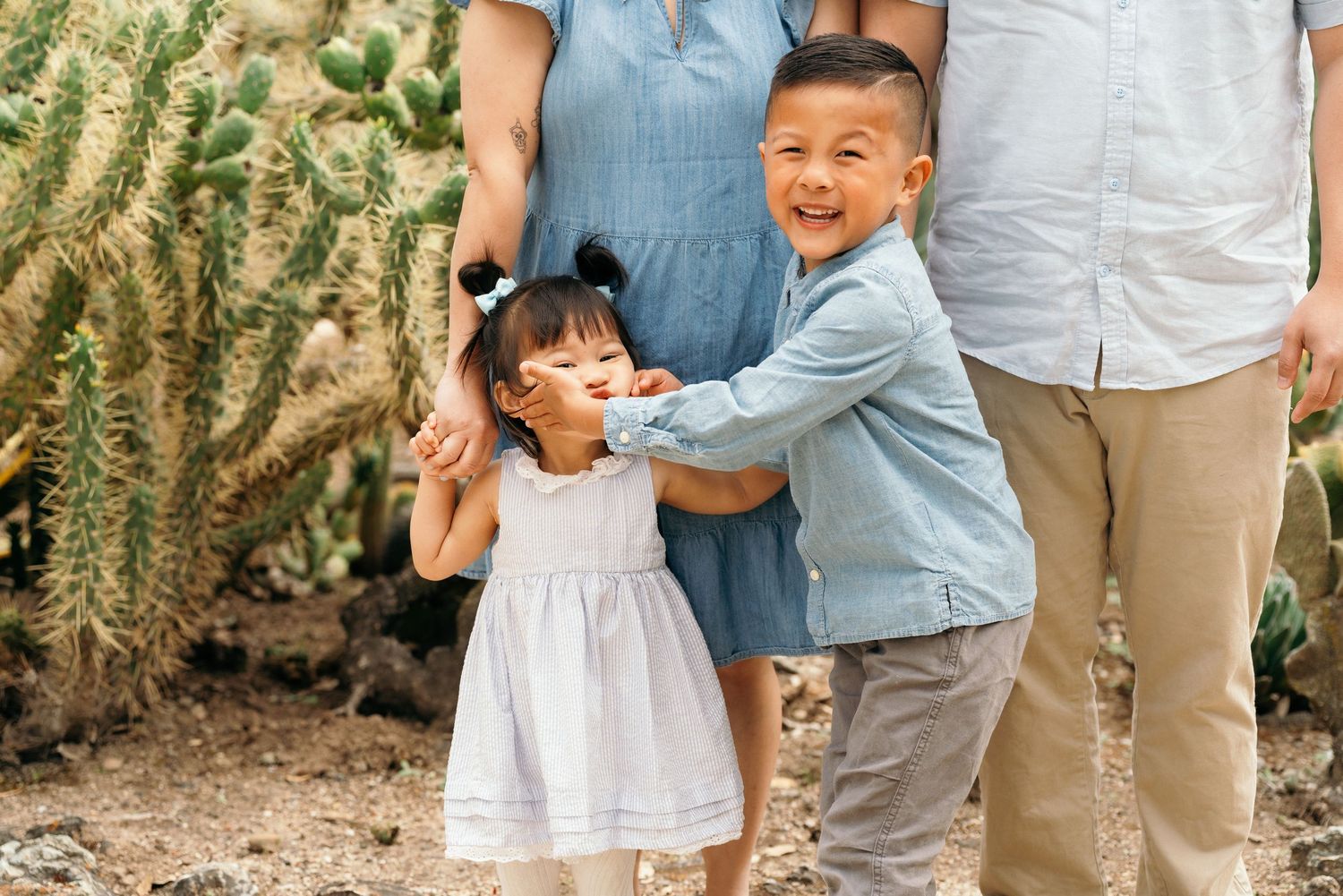 Asian family with two young children smiling outdoors near cactus plants, boy in blue shirt hugging little girl in white dress.