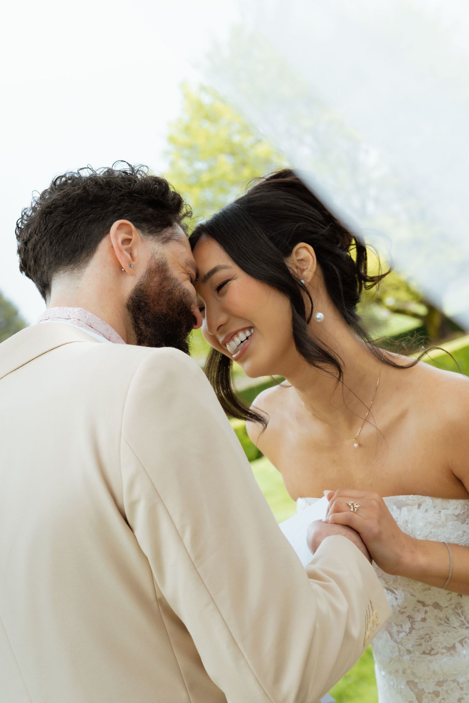 Laughing bride and groom in a tender embrace outdoors with lush green trees in the background.